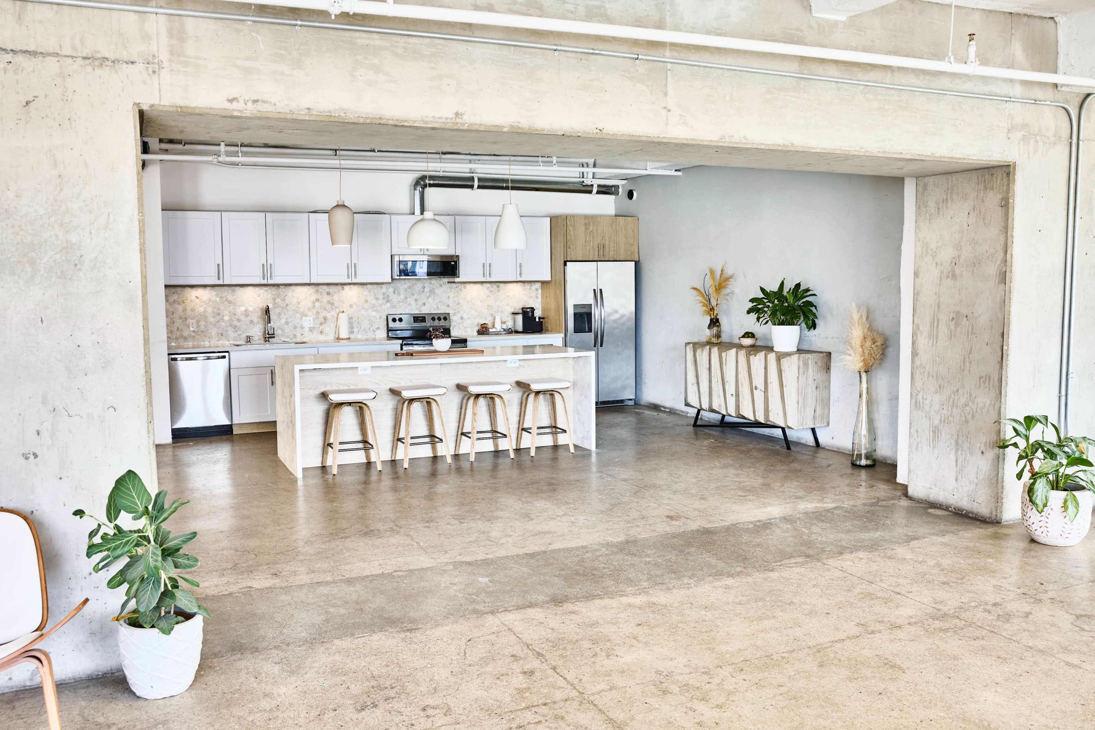 The image shows a modern kitchen and dining area with a minimalist design, featuring a large island with barstools and natural light streaming through an open space.