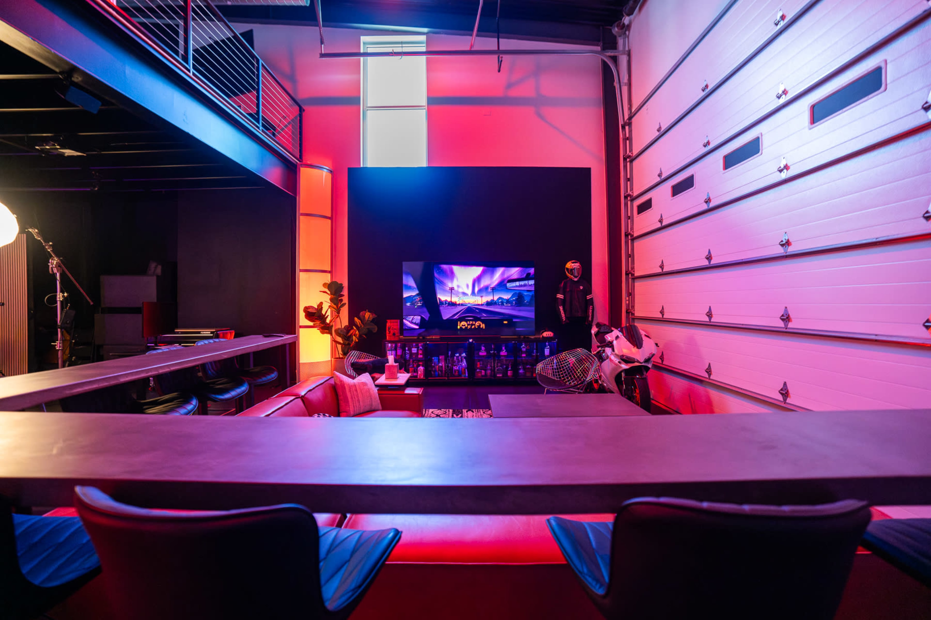 The image shows a modern, stylish interior with a large television, seating area, and colorful lighting, including a garage door on one side.
