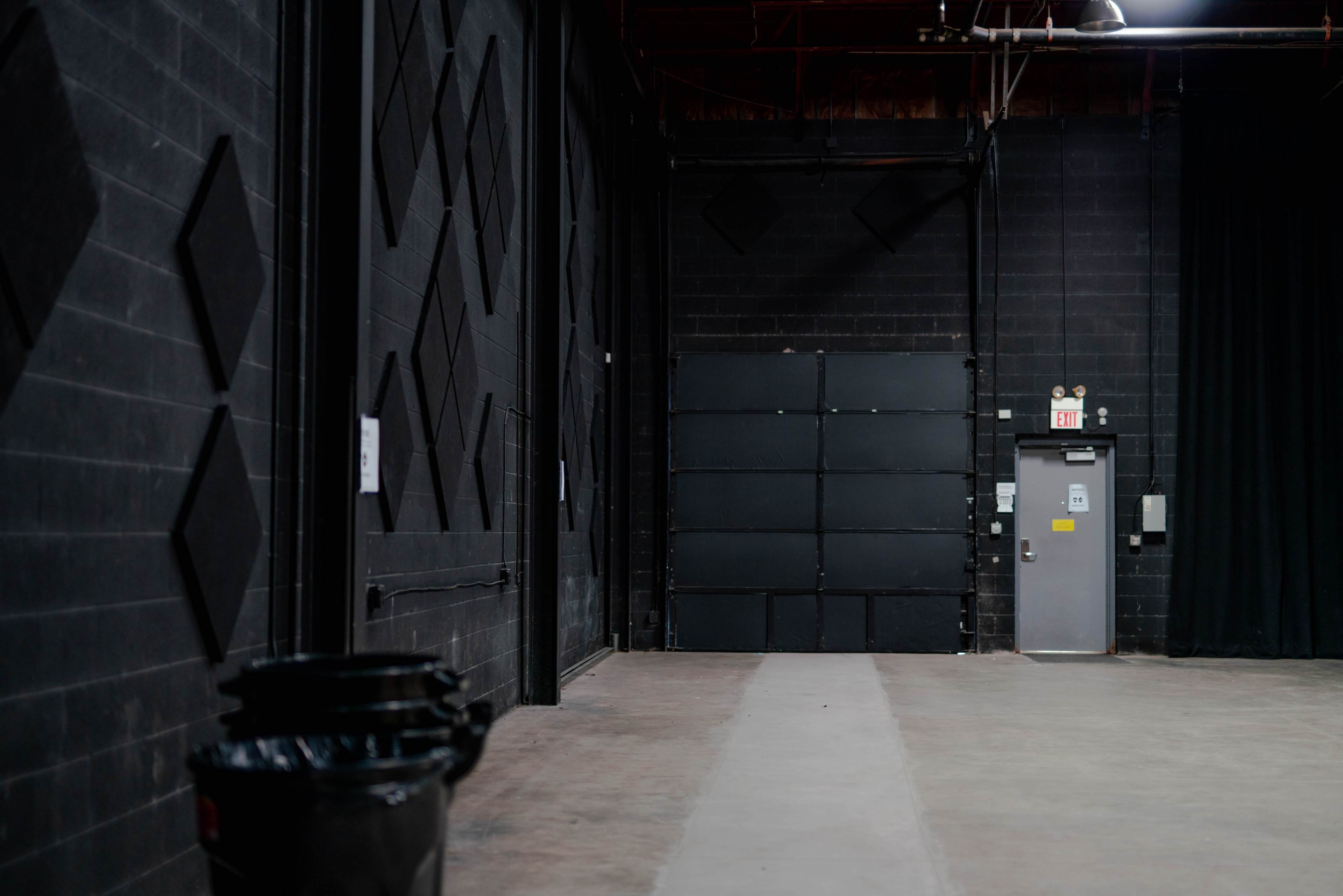 A long, empty hallway in a black-walled industrial space leads to a large closed door, with a fire exit and trash bins visible on the left.