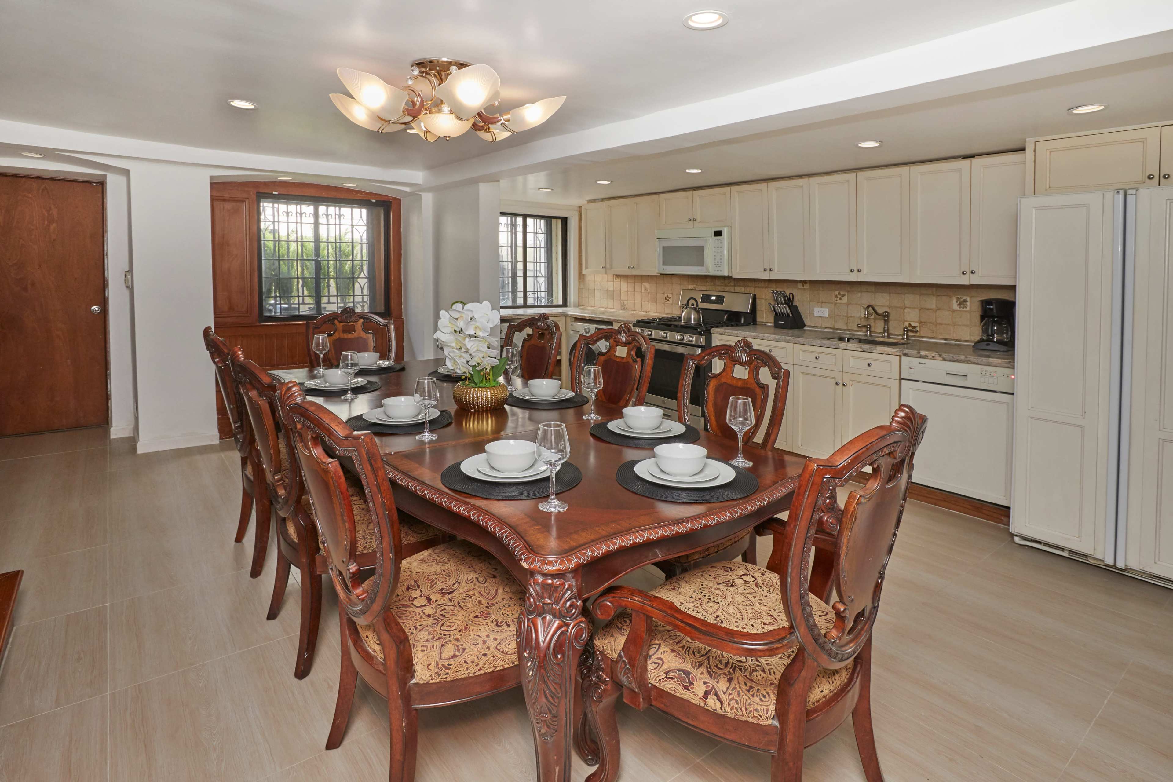 A dining room with a large wooden table set for eight, surrounded by ornate chairs, adjacent to a kitchen featuring light cabinetry and modern appliances.