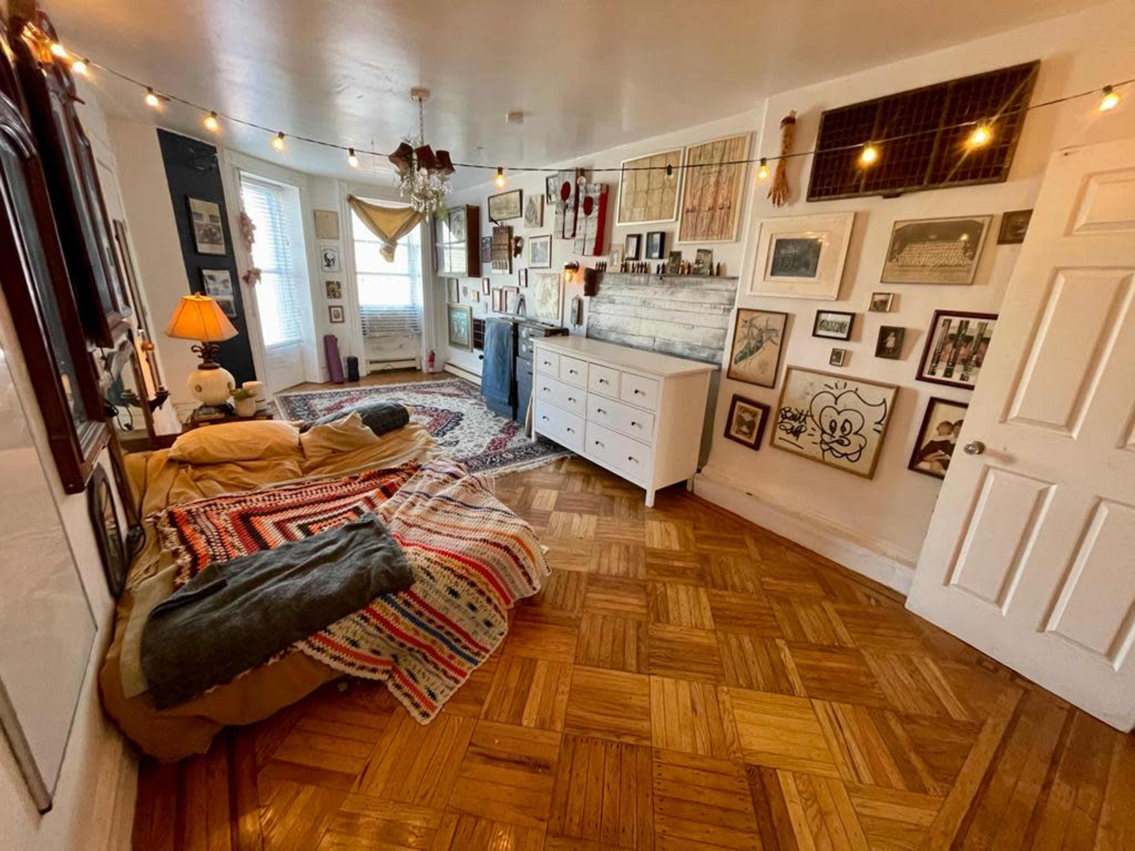 The image shows a cozy room with wooden flooring, a bed with a colorful blanket, and a wall adorned with various framed pictures and decorations.