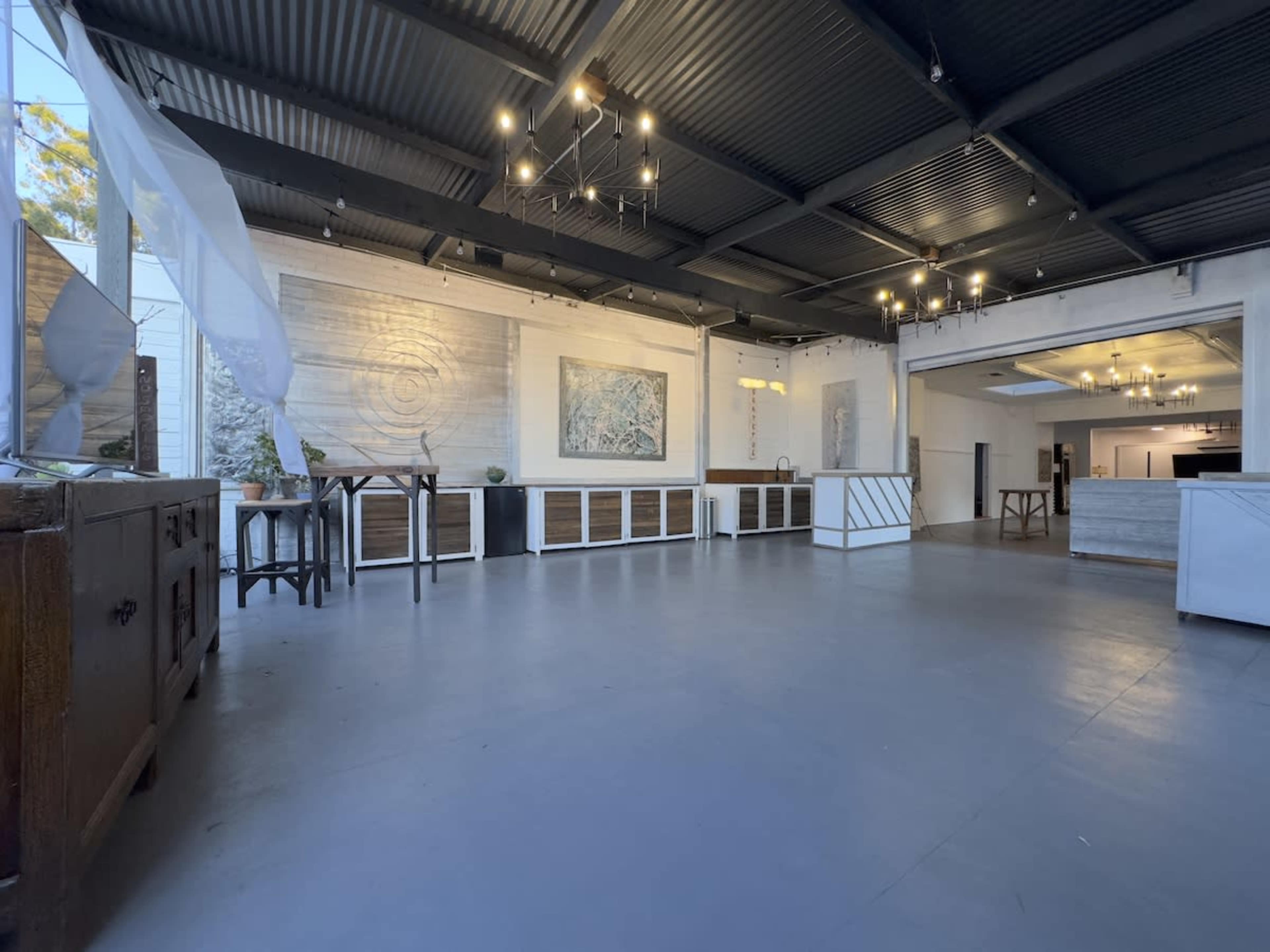 The image shows an empty event space with wooden furniture, exposed lighting fixtures, and large windows allowing natural light to enter.