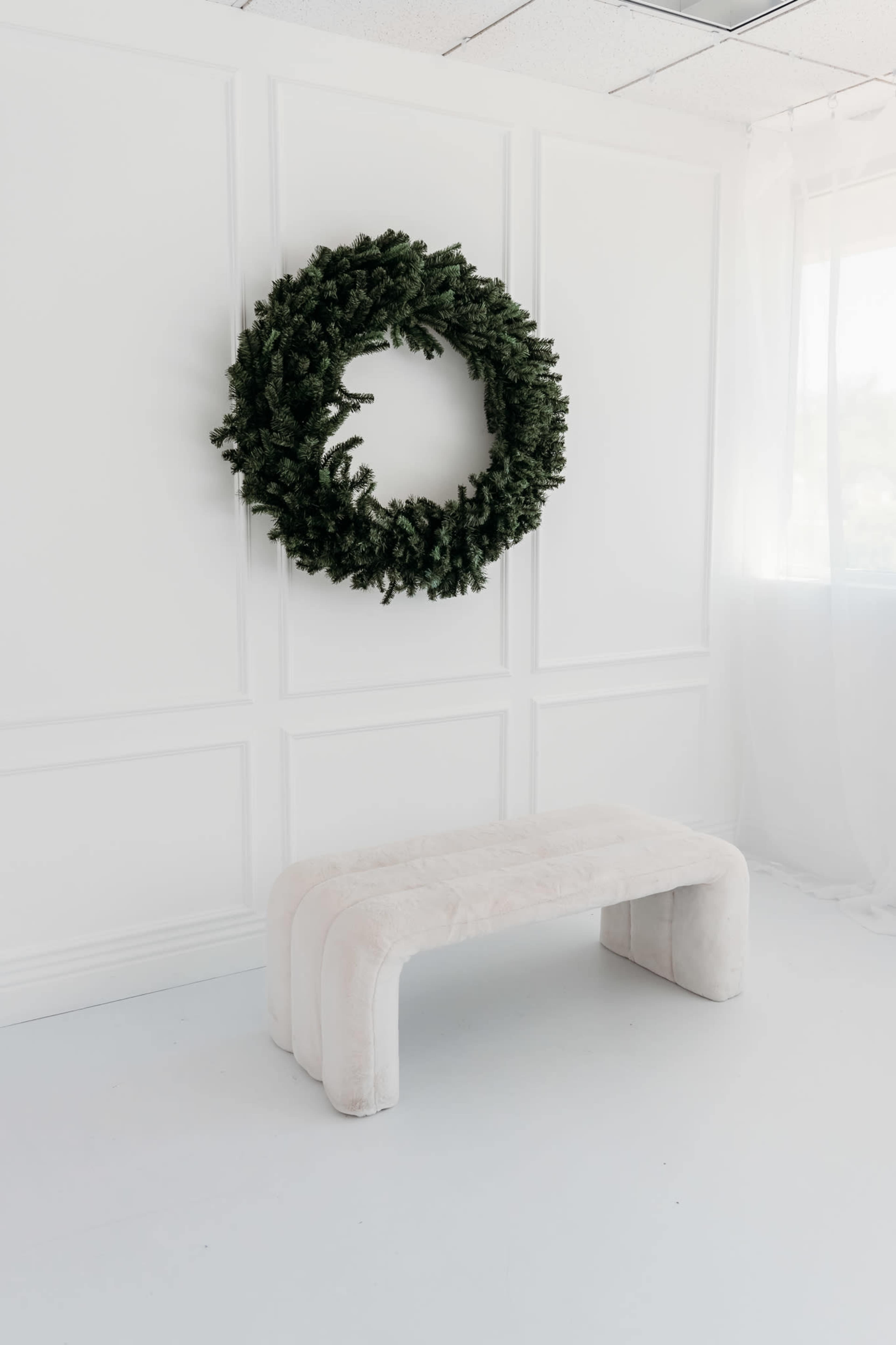 A large green wreath is mounted on a white wall above a soft, light-colored bench in a minimalistic room with a bright, airy atmosphere.