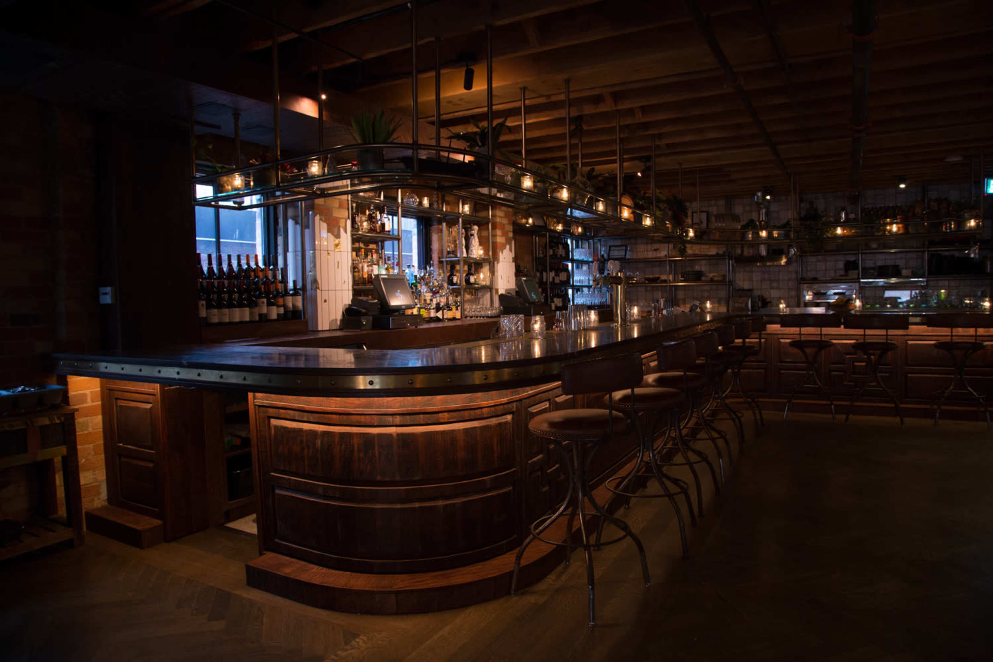 The image shows a dimly lit bar with a curved wooden counter and metal accents, featuring several empty stools and shelves lined with bottles in the background.