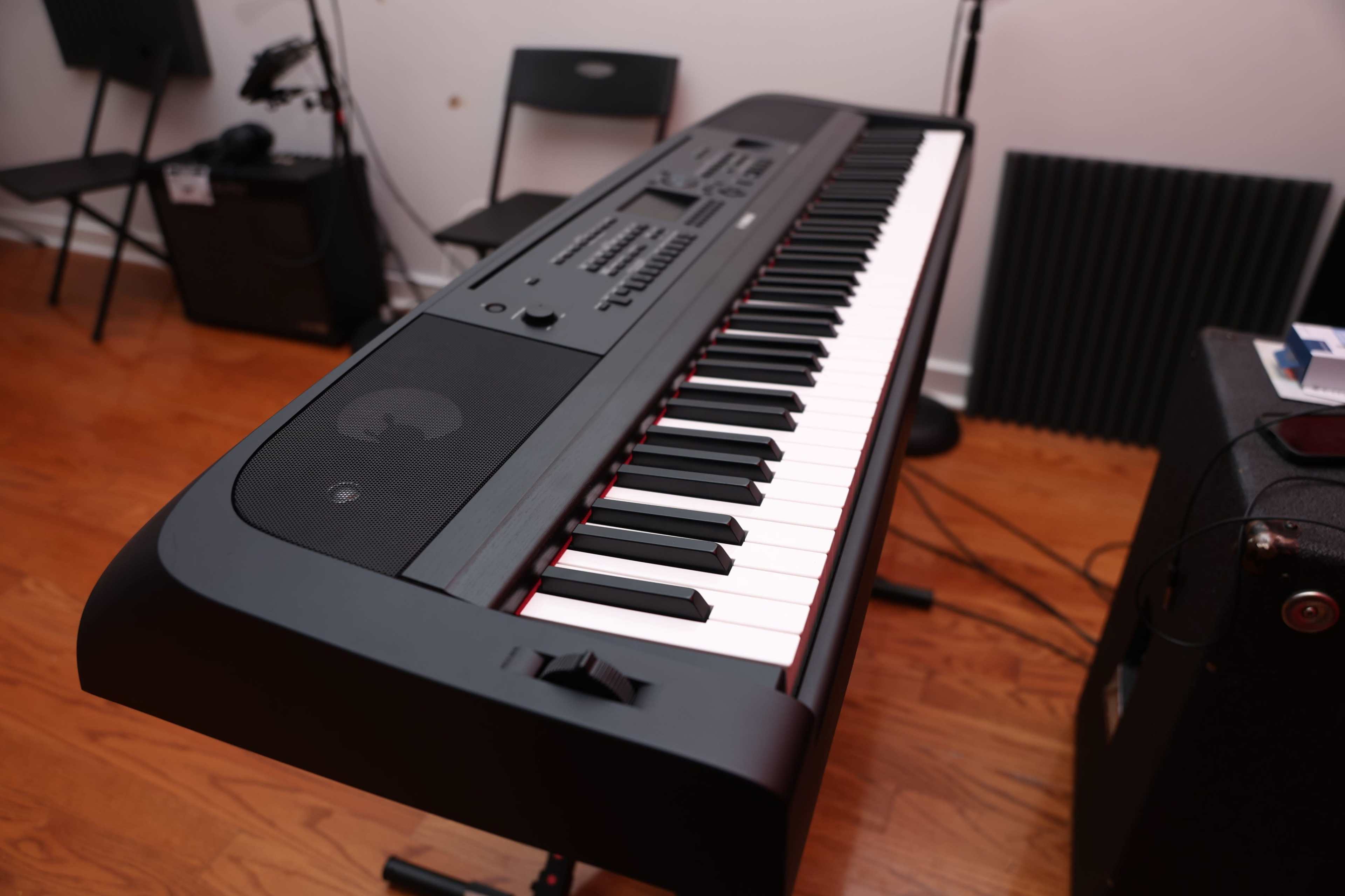 The image features a close-up of a digital piano with black keys and a sleek design, positioned in a room with wooden flooring and seating in the background.