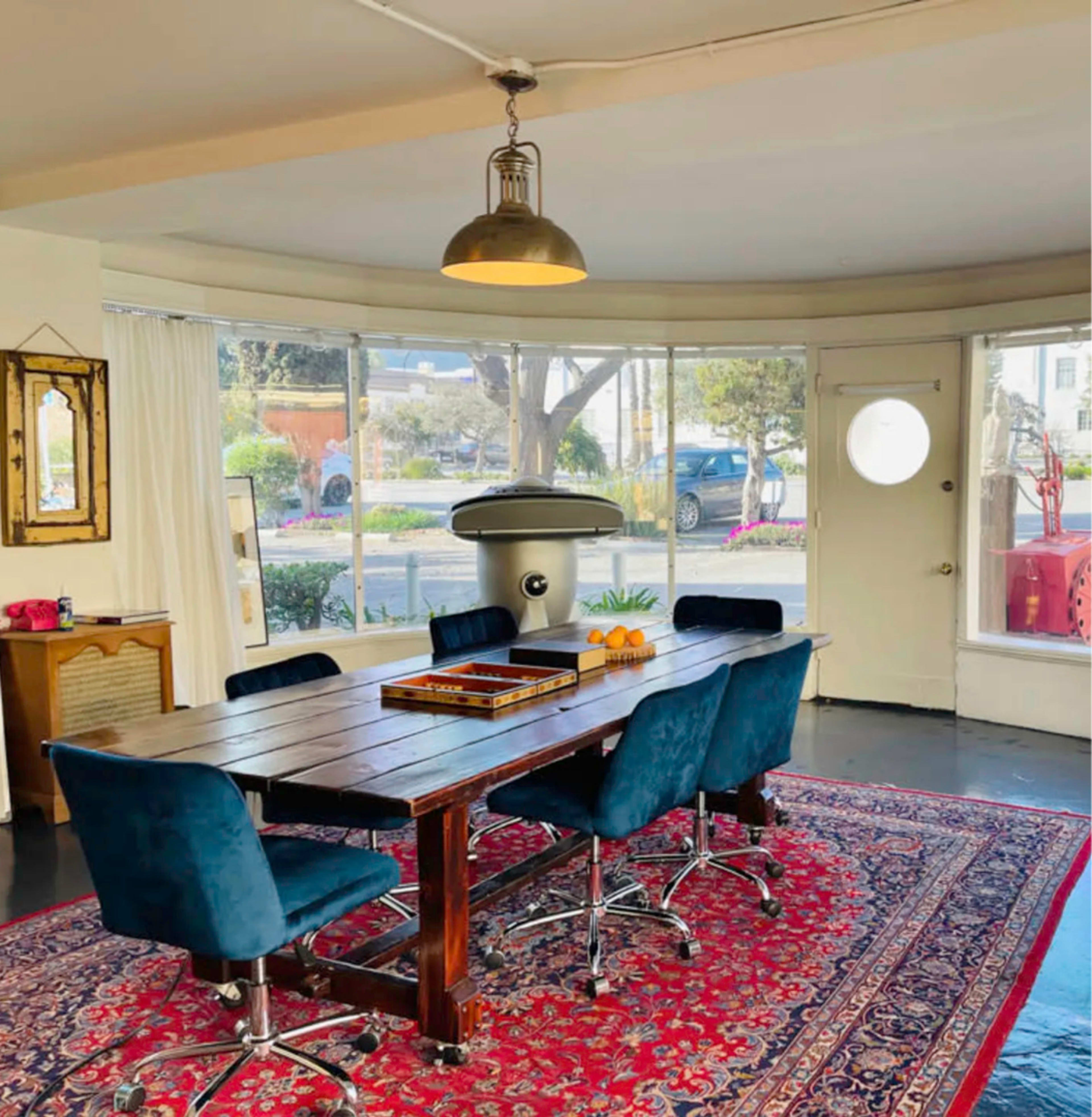A spacious dining area features a wooden table surrounded by blue chairs, with a red patterned rug beneath and windows allowing natural light to enter.