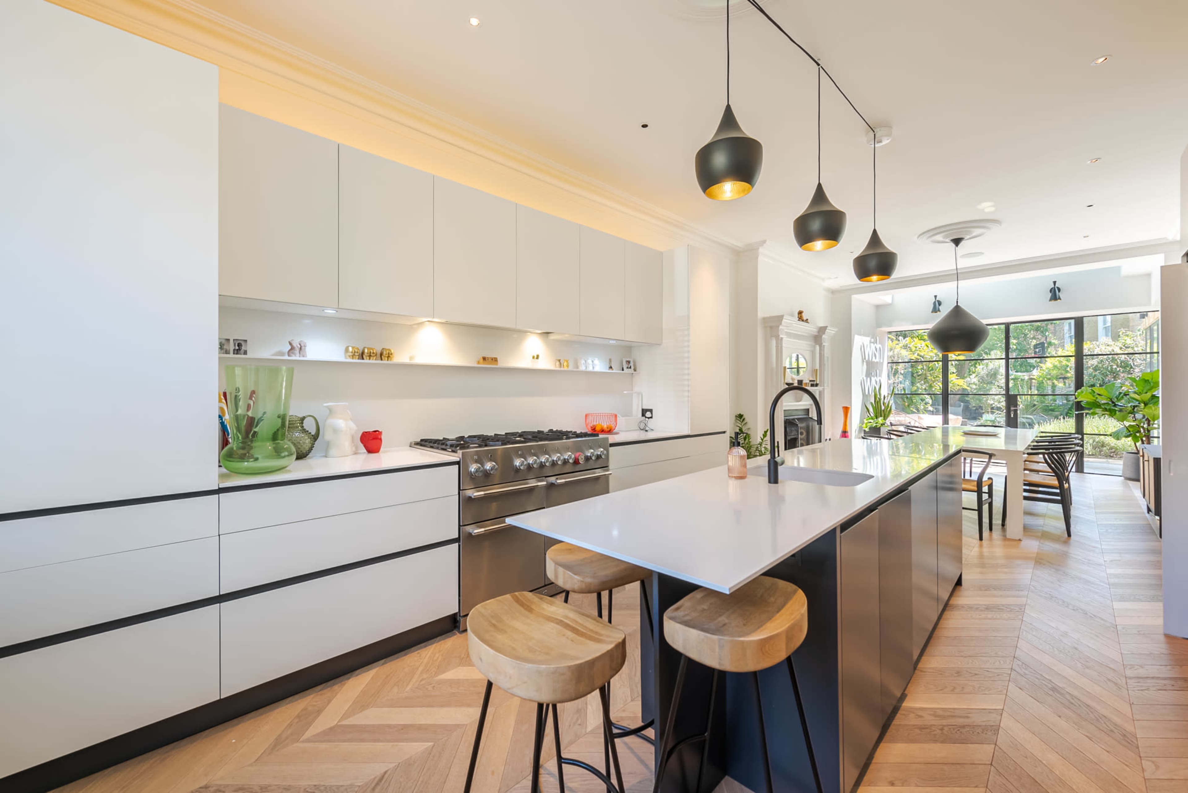 A modern kitchen features a central island with bar stools, sleek cabinetry, stainless steel appliances, and large windows allowing natural light.