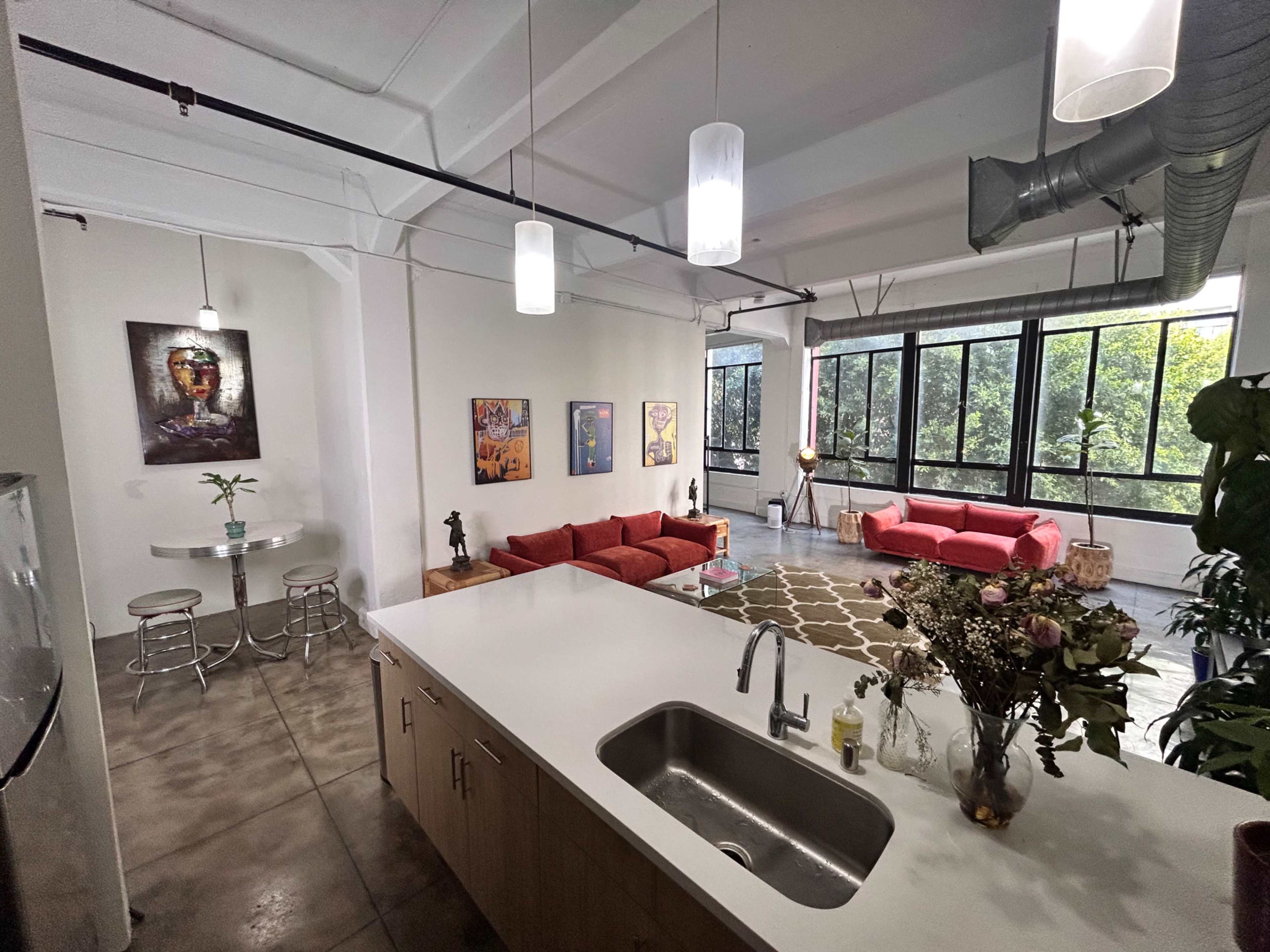 The image shows a spacious loft-style apartment with modern furnishings, including a kitchen island, red sofas, and large windows allowing natural light.