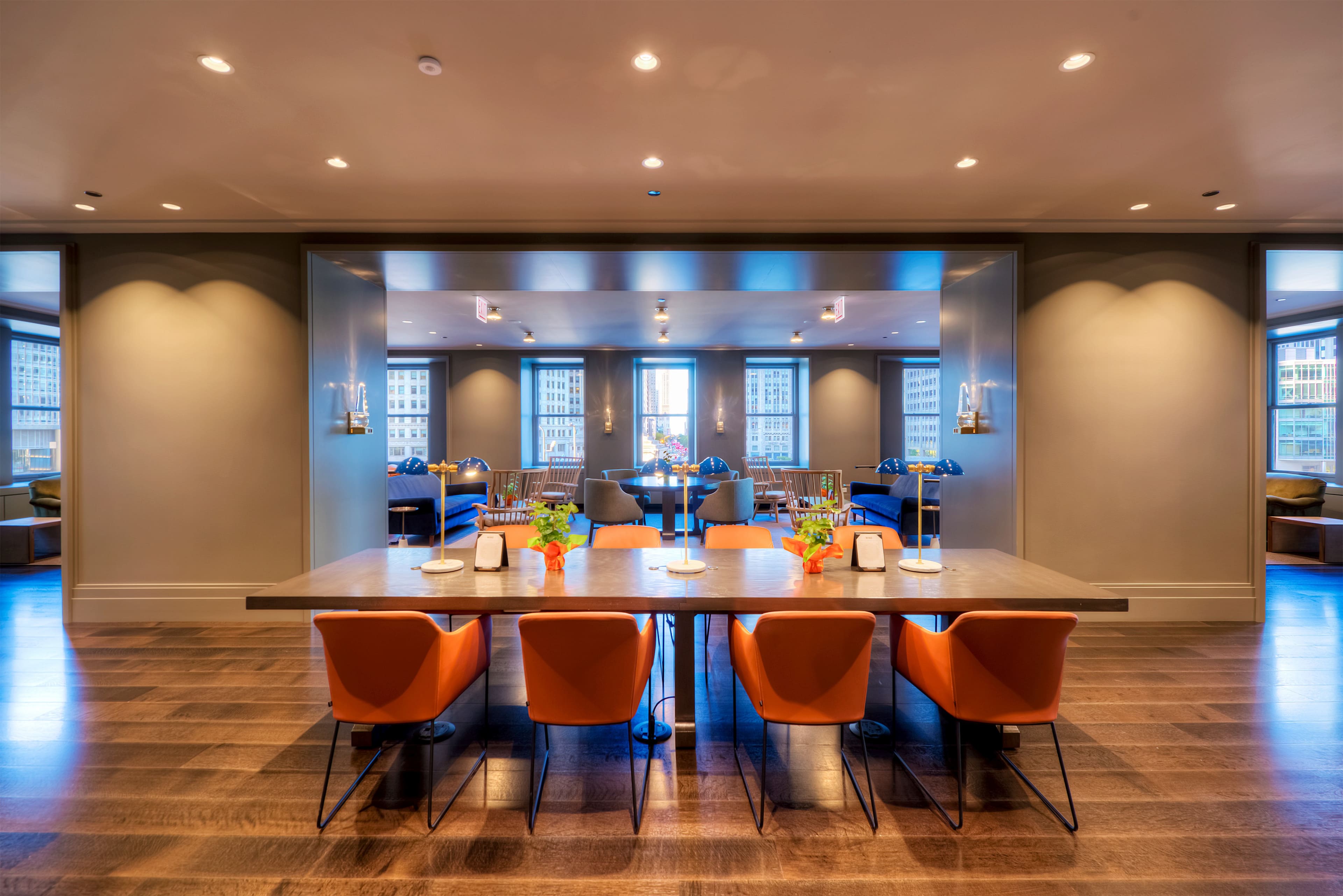 A modern dining area with a large table surrounded by orange chairs, leading into a well-lit lounge area with large windows.