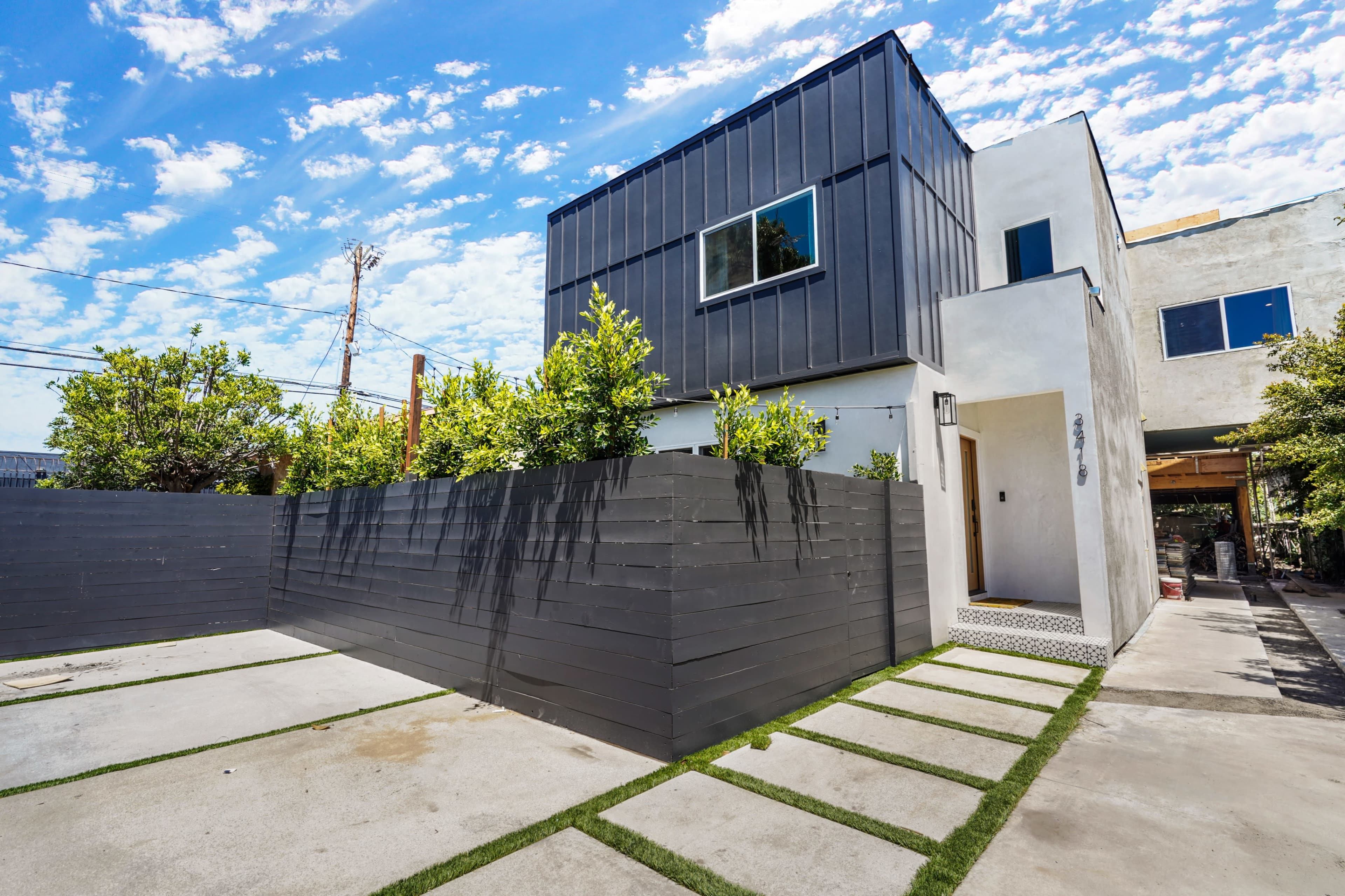 A modern two-story house features a mix of metal and stucco exteriors, surrounded by a gray privacy fence and green landscaping elements in the front yard.