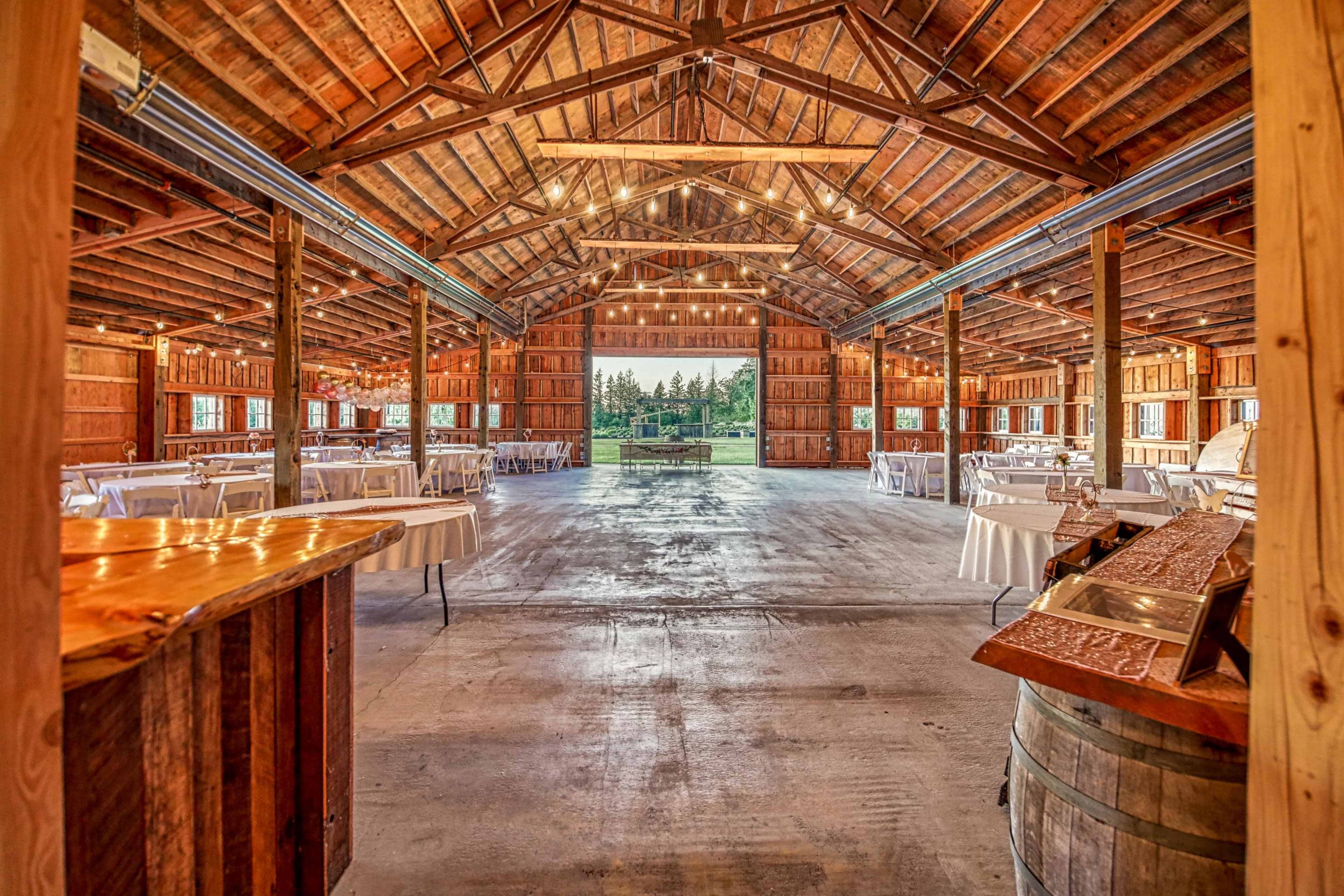 The interior of a spacious barn venue features wooden beams, table setups, and large openings that lead to an outdoor view.