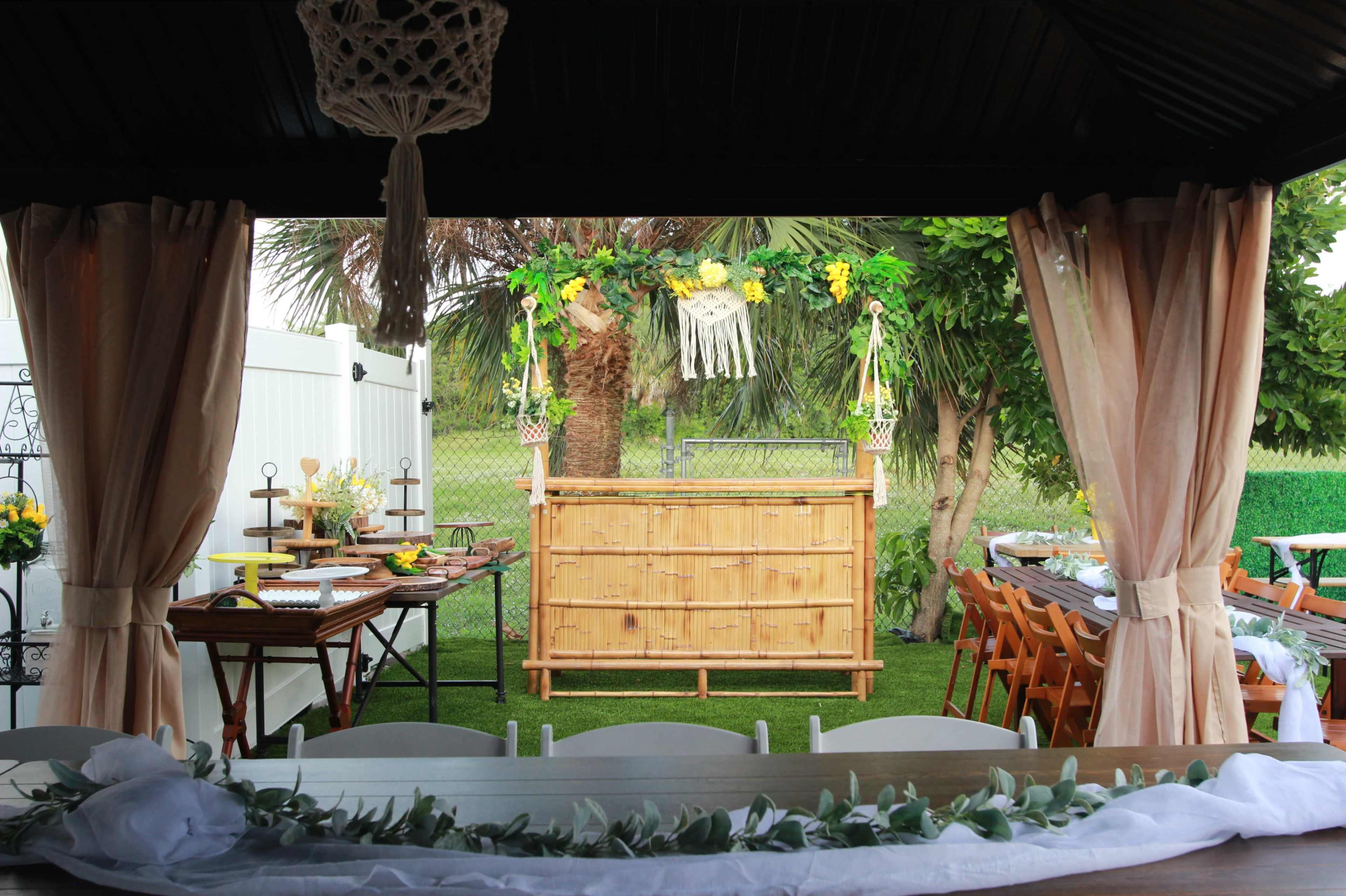 A bamboo bar adorned with decorations and surrounded by tables and chairs is set up under a shelter for an outdoor gathering.