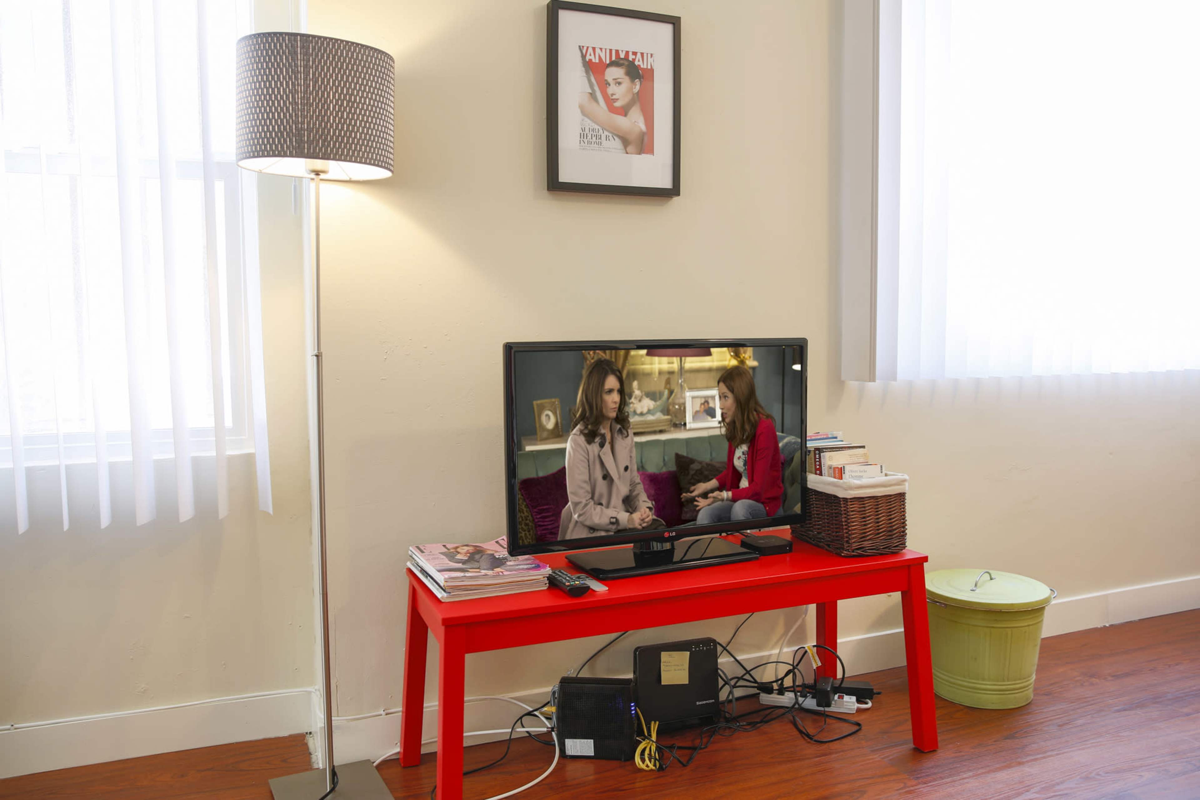 A red table holds a television displaying two women conversing, surrounded by a lamp, magazines, and a basket, against a wall with a framed magazine cover.