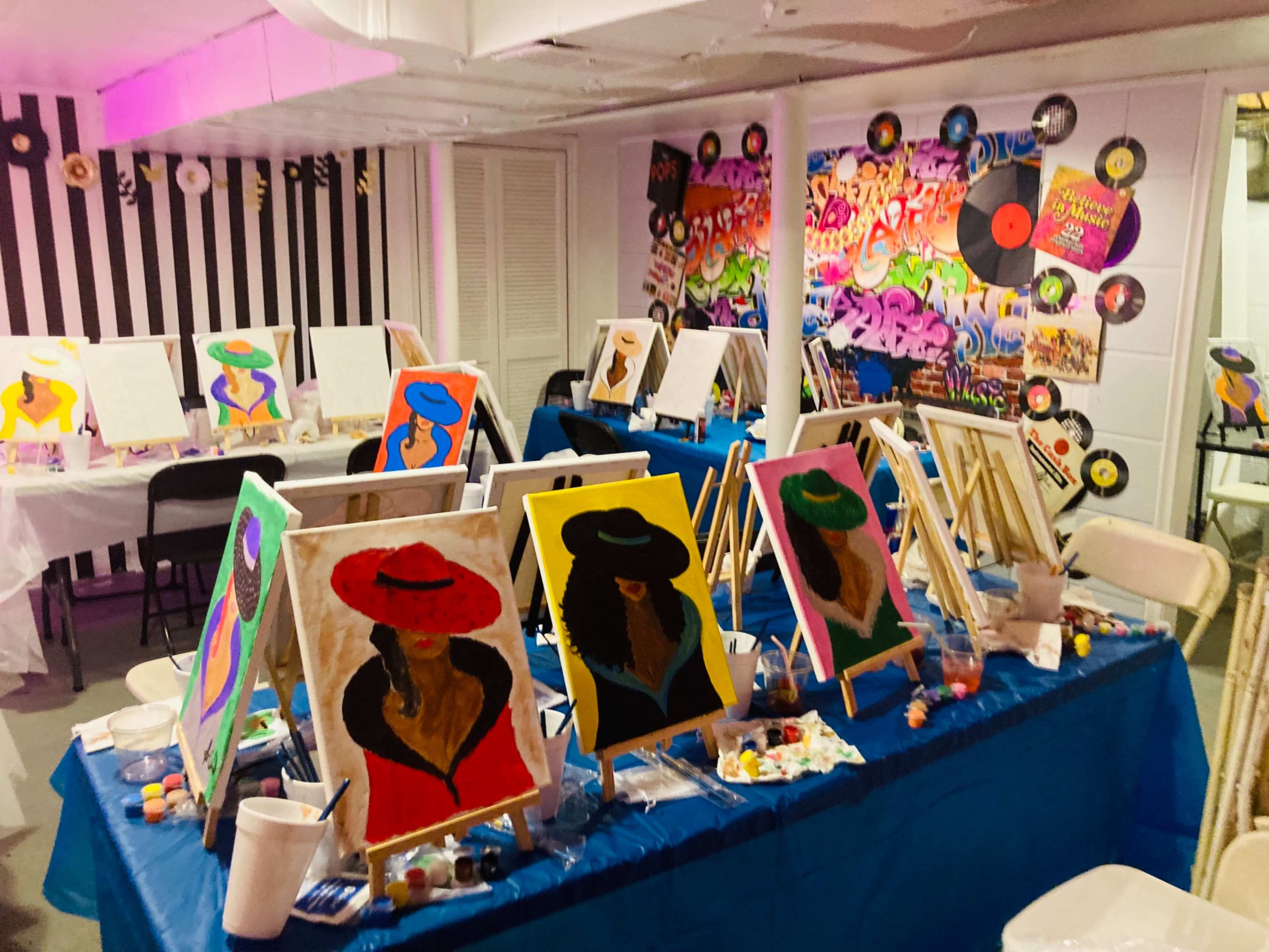 A brightly decorated art studio with multiple easels displaying paintings of women wearing hats, surrounded by painting supplies and a colorful graffiti-style mural on the wall.