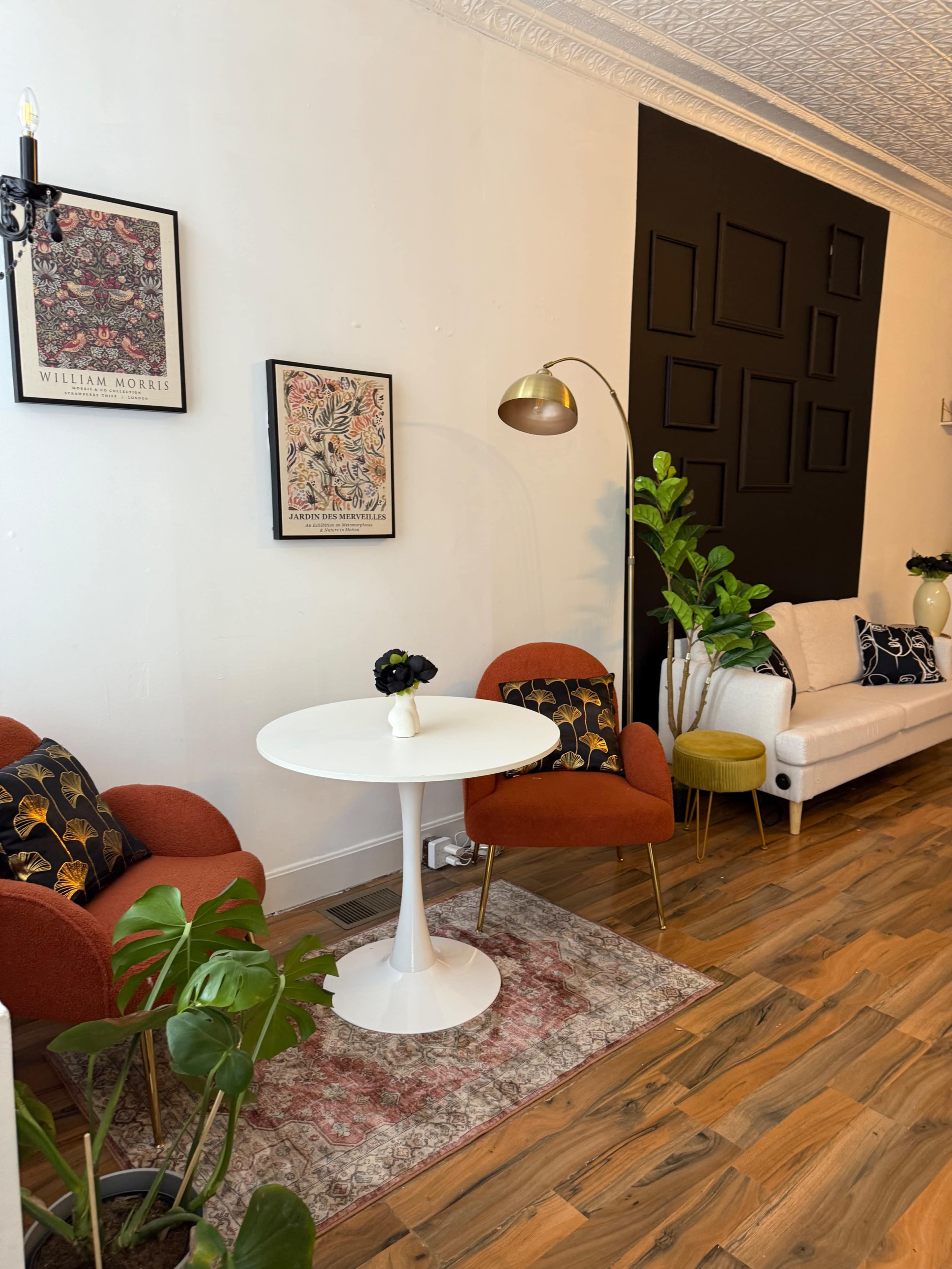 The image shows a stylish living room with a white sofa, orange chairs, a round table, decorative plants, and artwork on the walls.