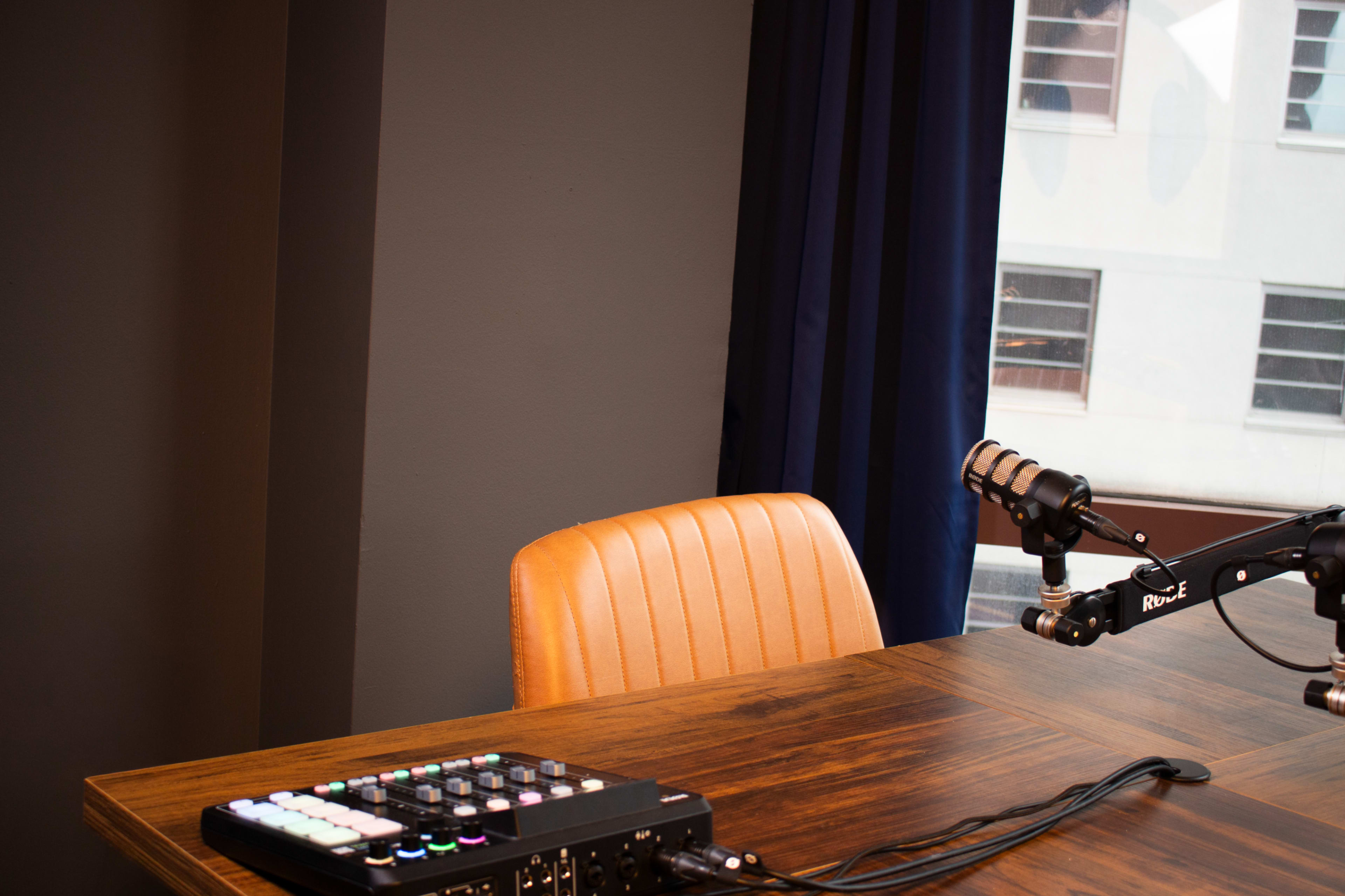 The image shows a recording studio setup with a wooden table, an empty orange chair, a microphone on an arm, and an audio mixer next to it.