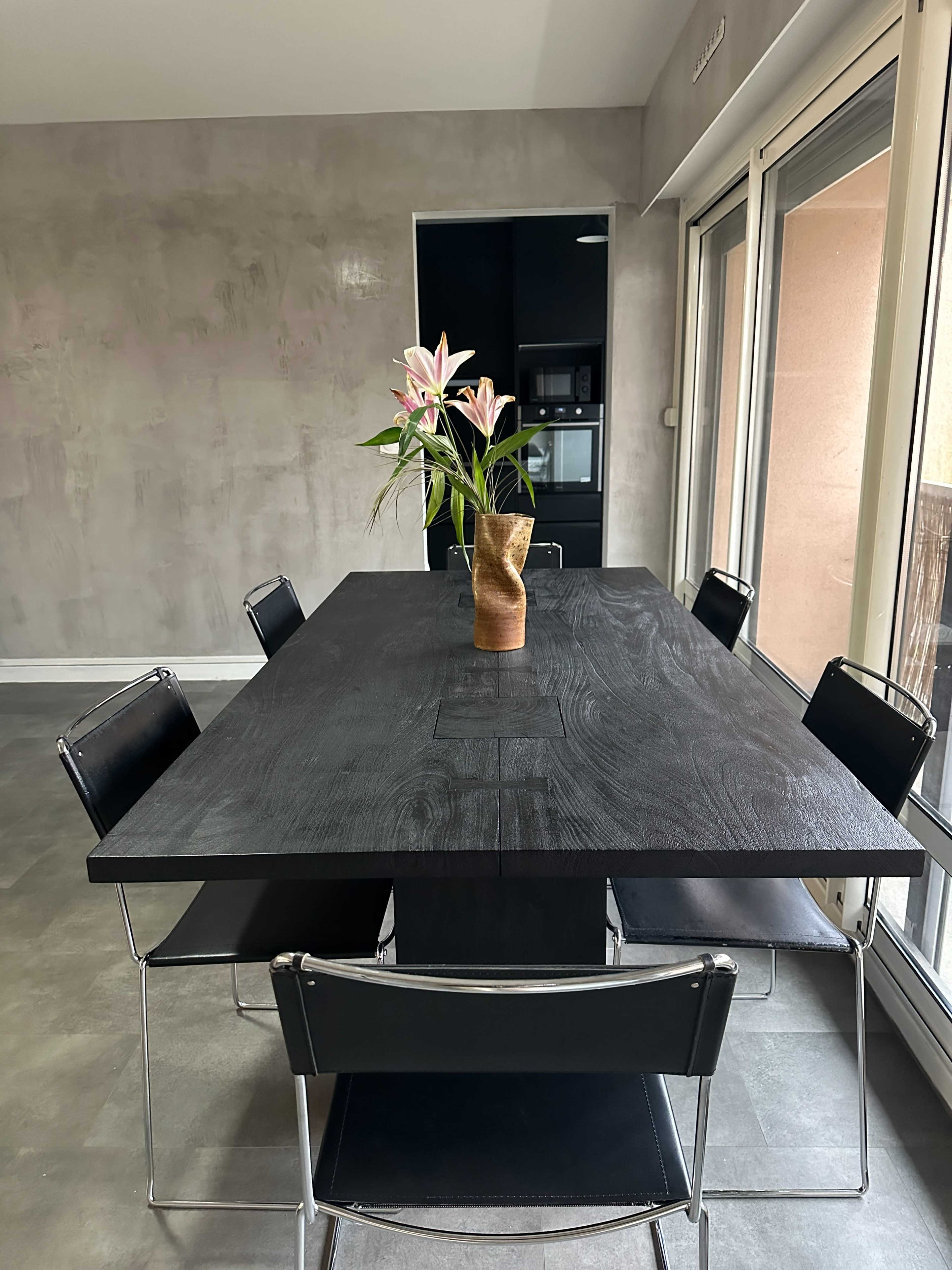 A dark wooden table with a vase of flowers is positioned in a modern dining area with light gray walls and large windows.
