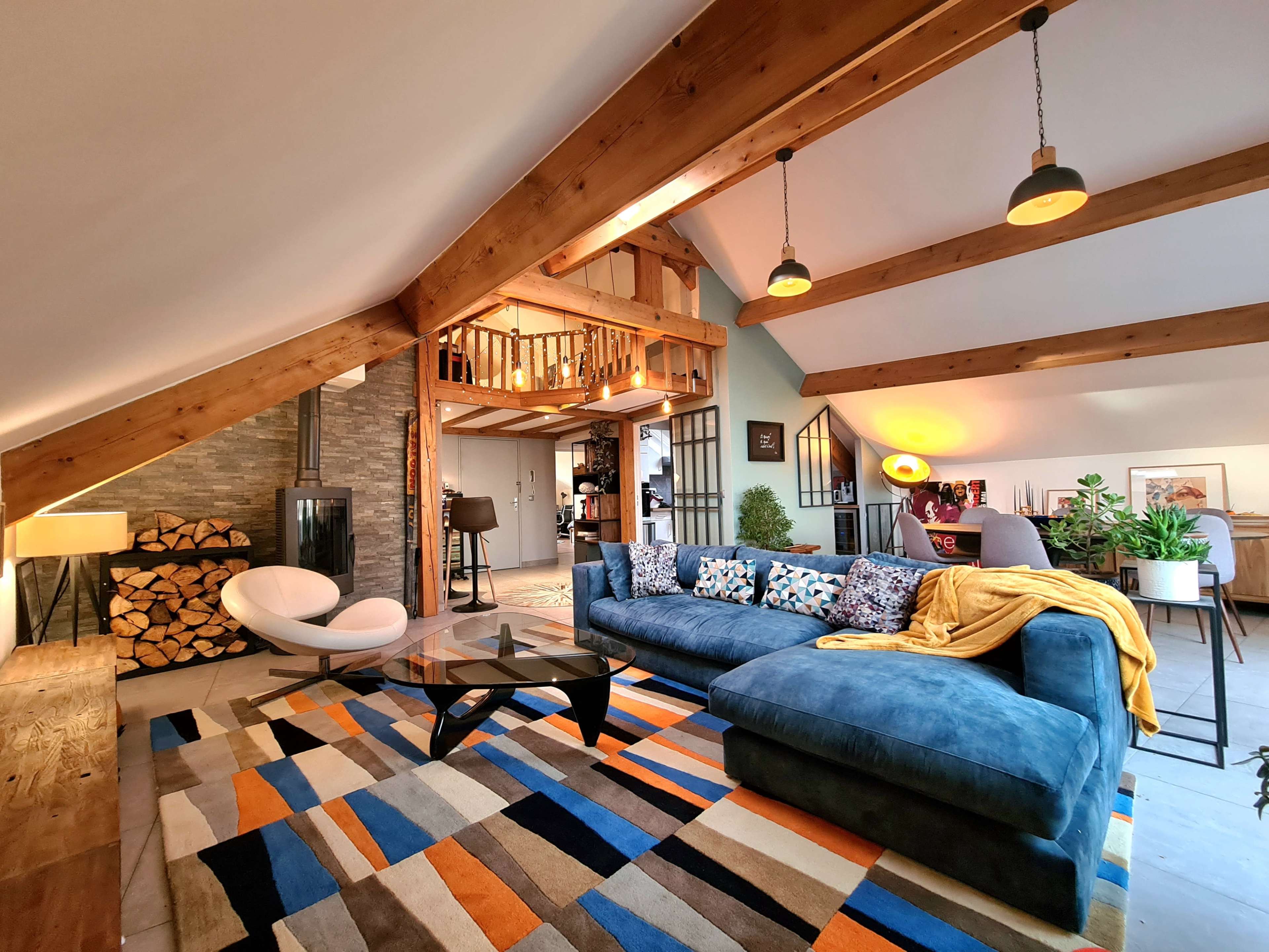 A well-furnished living room with a large blue sectional sofa, a coffee table, a colorful area rug, and exposed wooden beams on the ceiling.