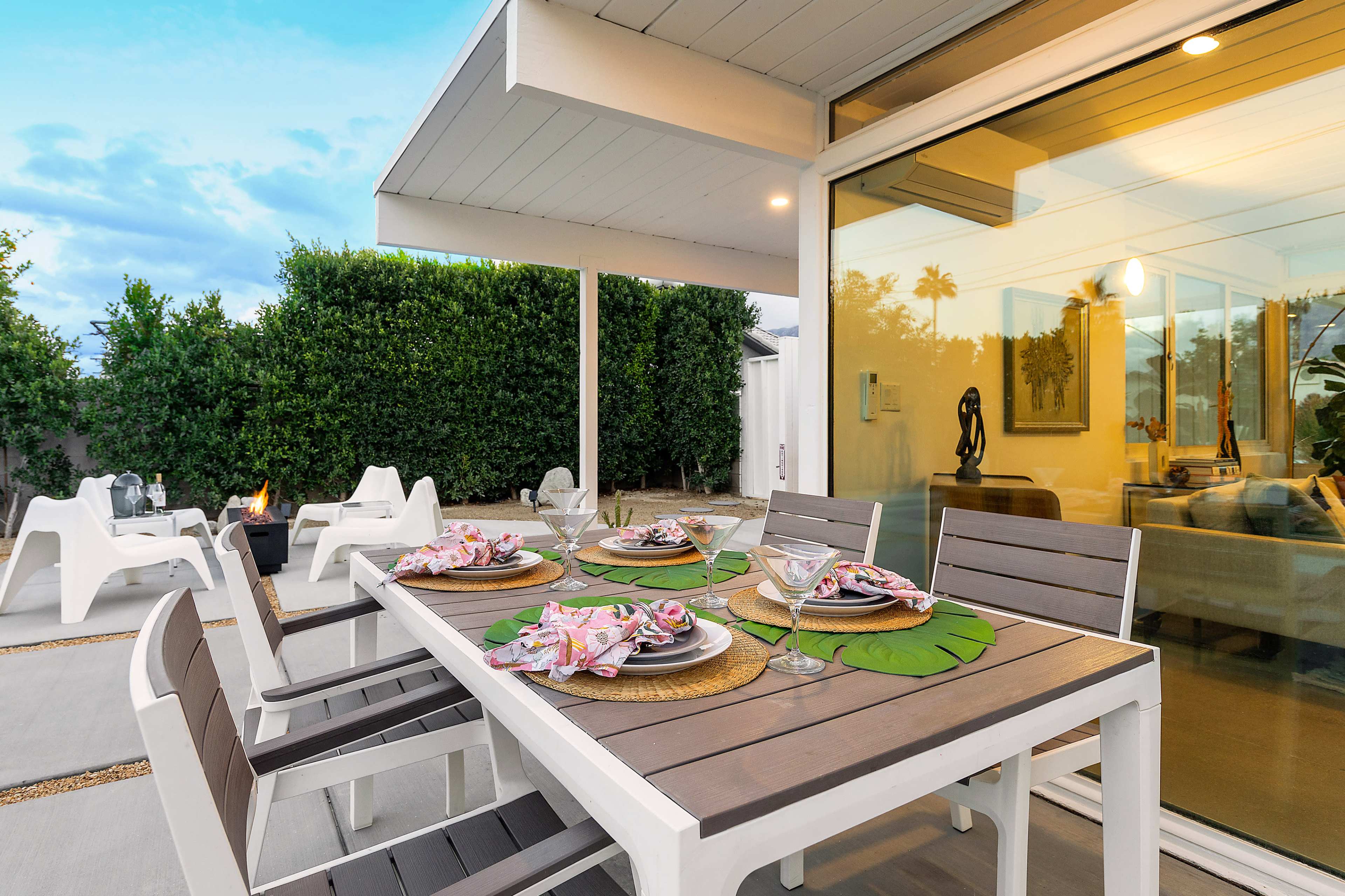 A modern outdoor dining area with a table set for four, featuring plates and glassware, and a view of a landscaped garden in the background.