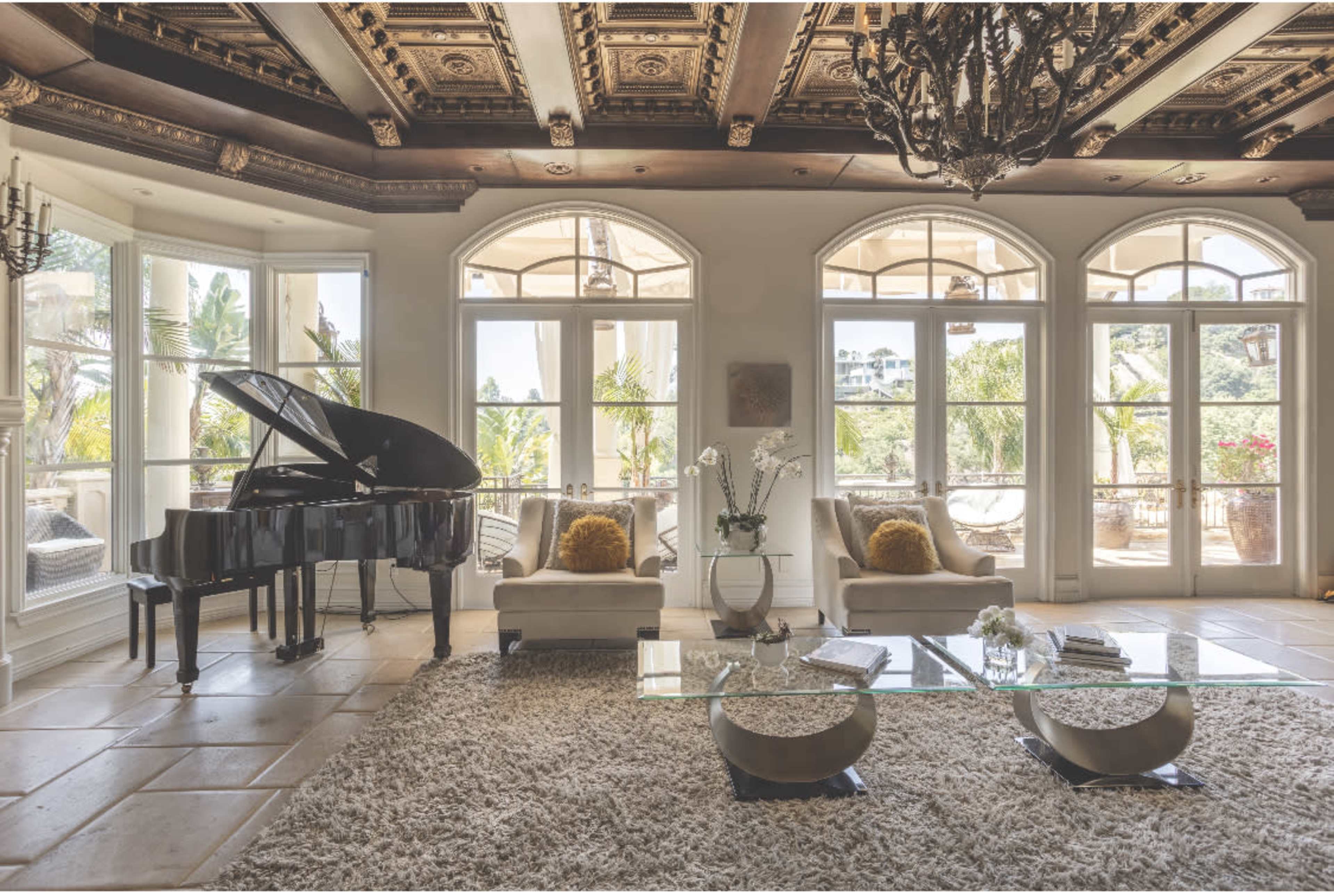 The image shows a spacious living room featuring a grand piano, two armchairs, a glass coffee table, and large windows that provide natural light and a view of outdoor greenery.