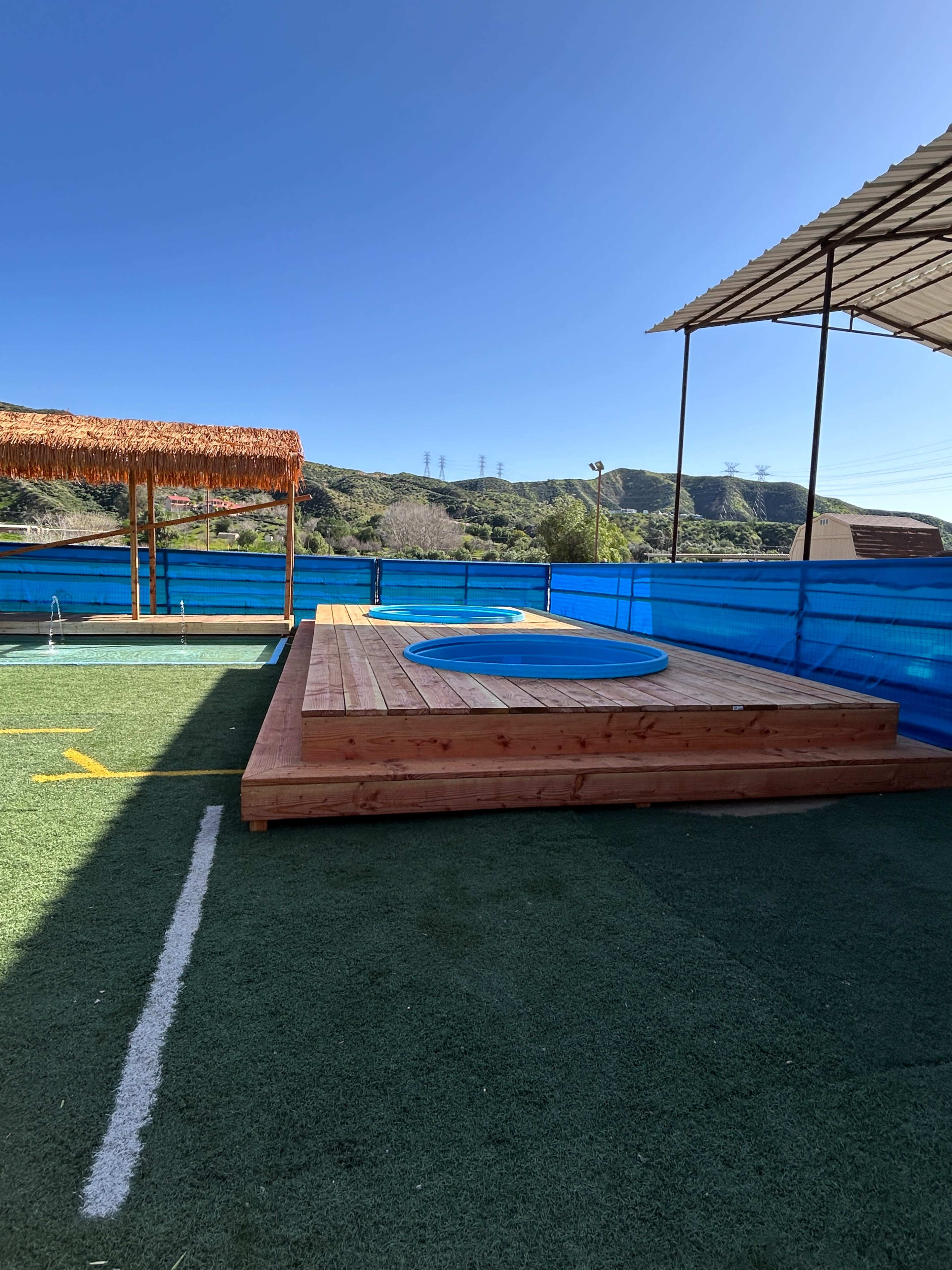The image shows a wooden deck with a circular blue pool, surrounded by green artificial grass and a blue barrier, under a clear blue sky.