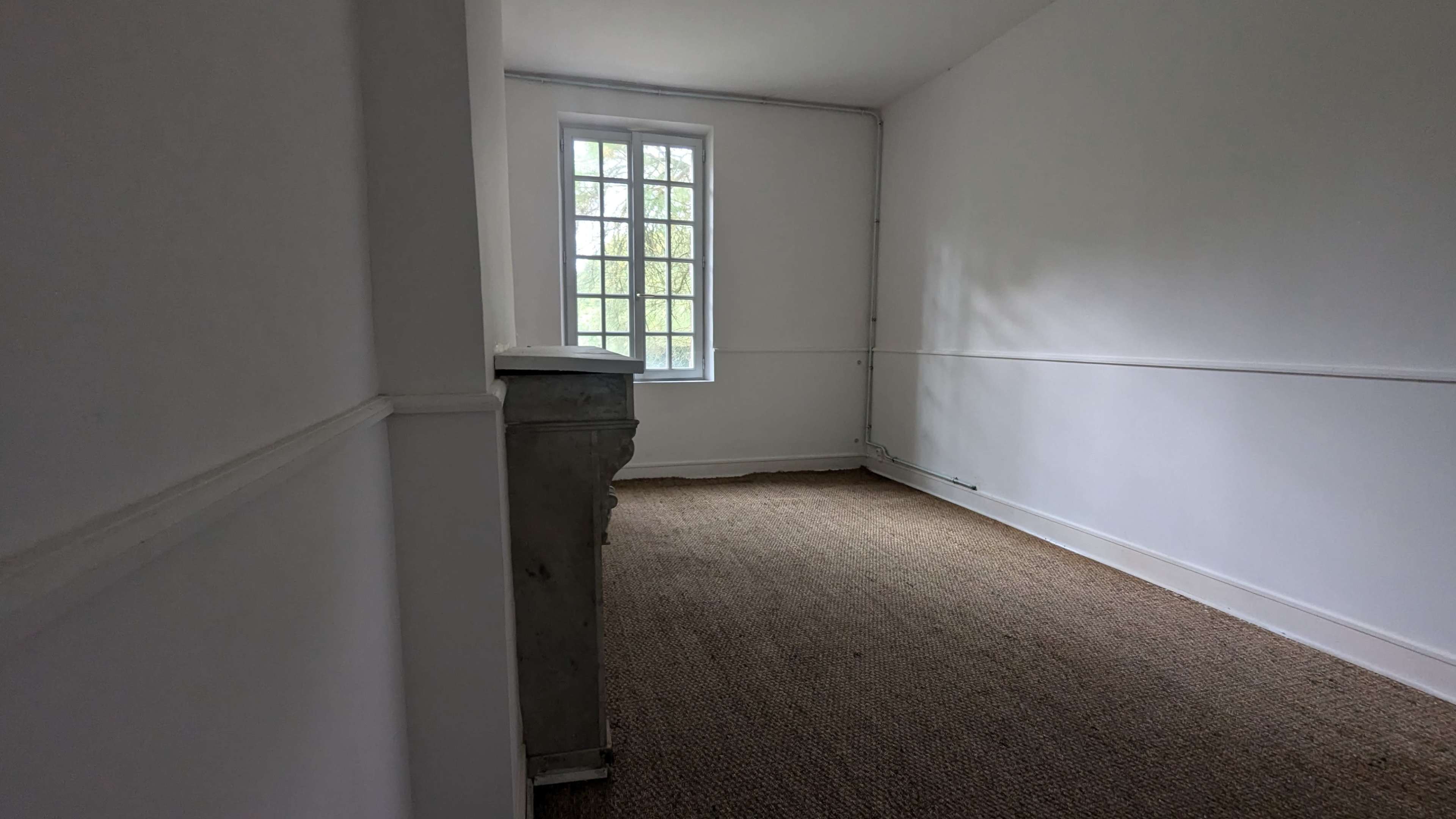 The image shows a sparsely furnished, empty room with a carpeted floor and a large window on one wall.