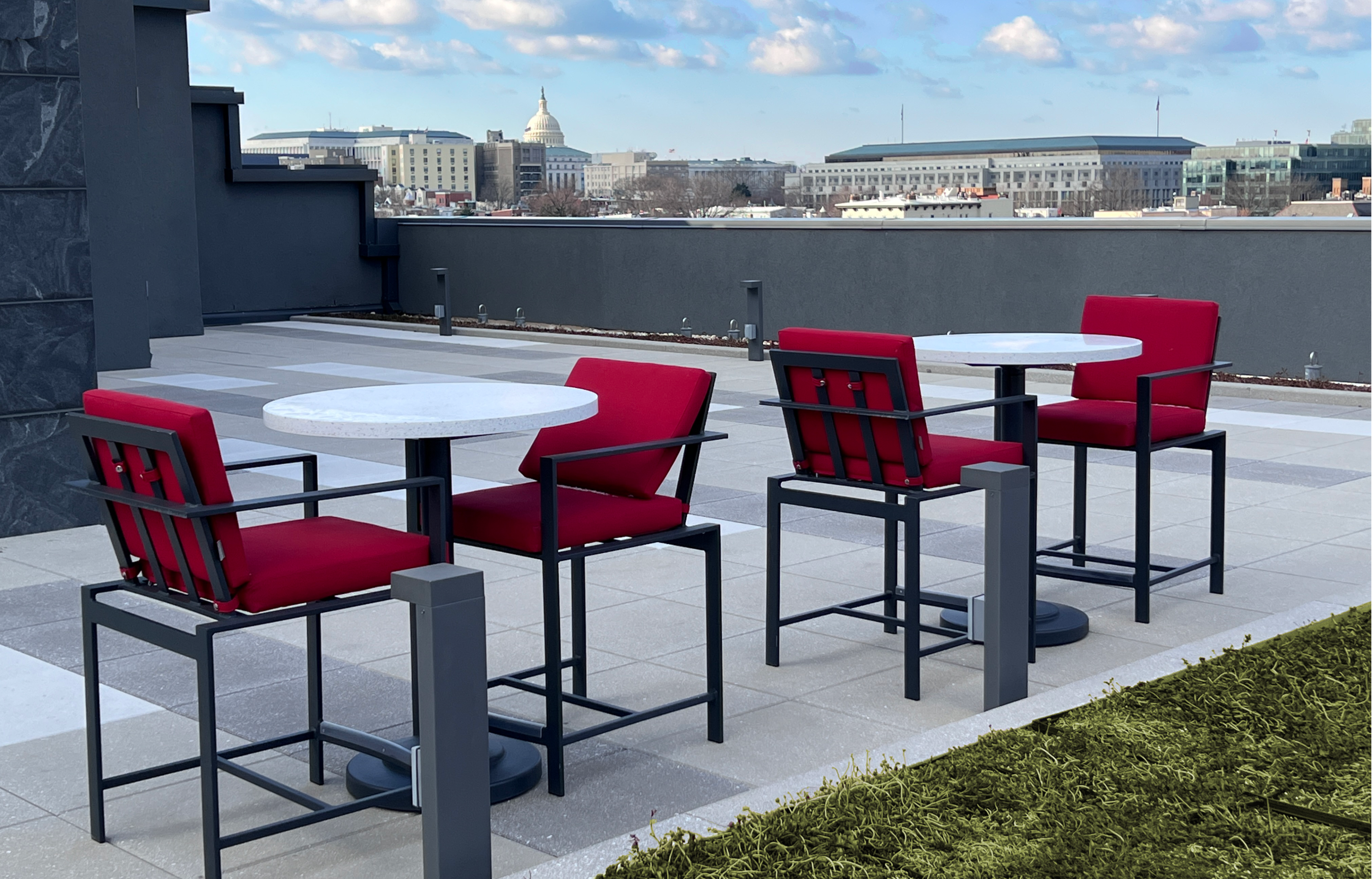 Two modern outdoor tables with red chairs on a rooftop terrace overlooking city buildings and a dome structure in the background.