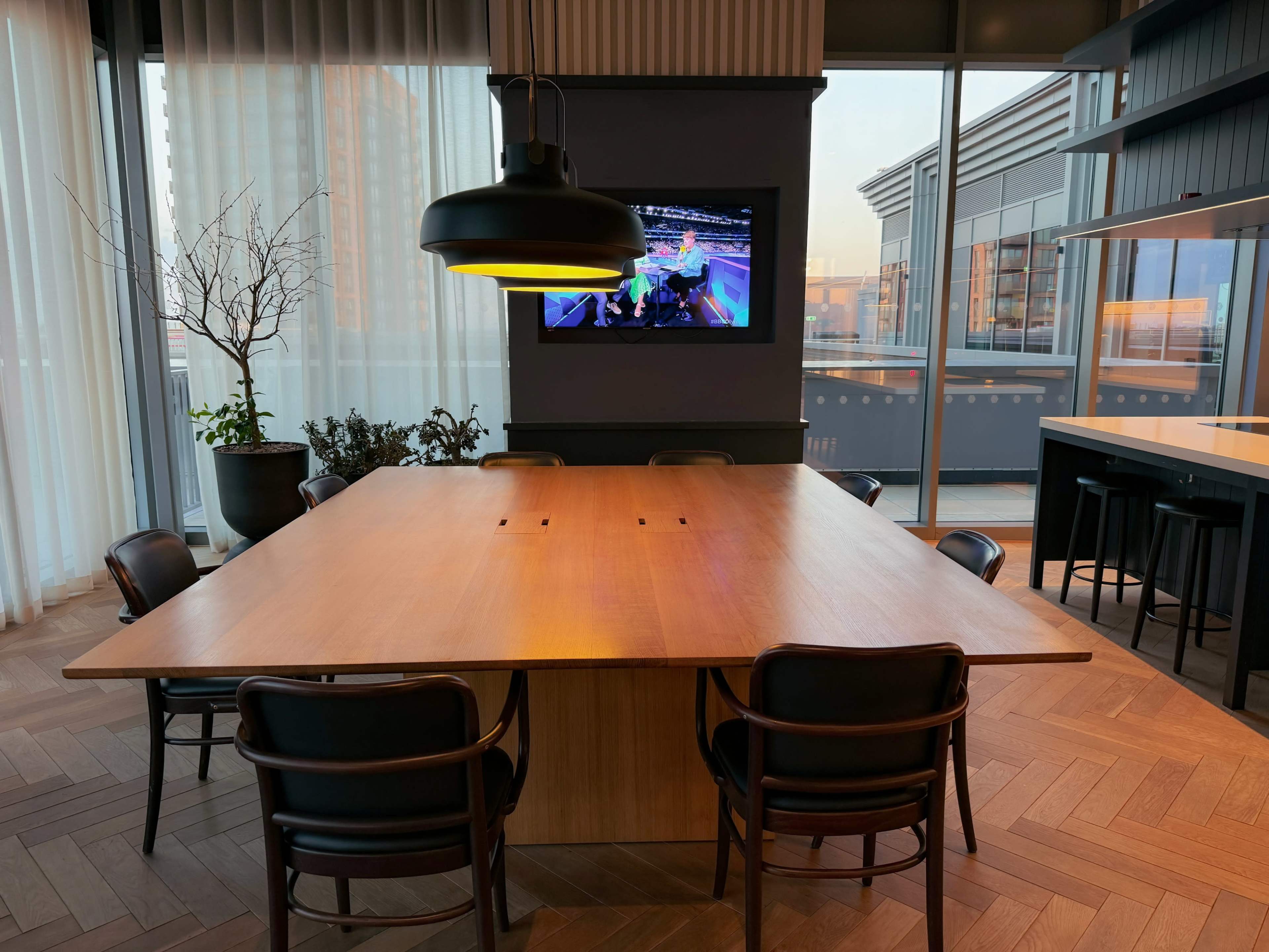 A large wooden conference table is surrounded by black chairs, with a television mounted on the wall displaying a colorful image and large windows allowing natural light to fill the space.