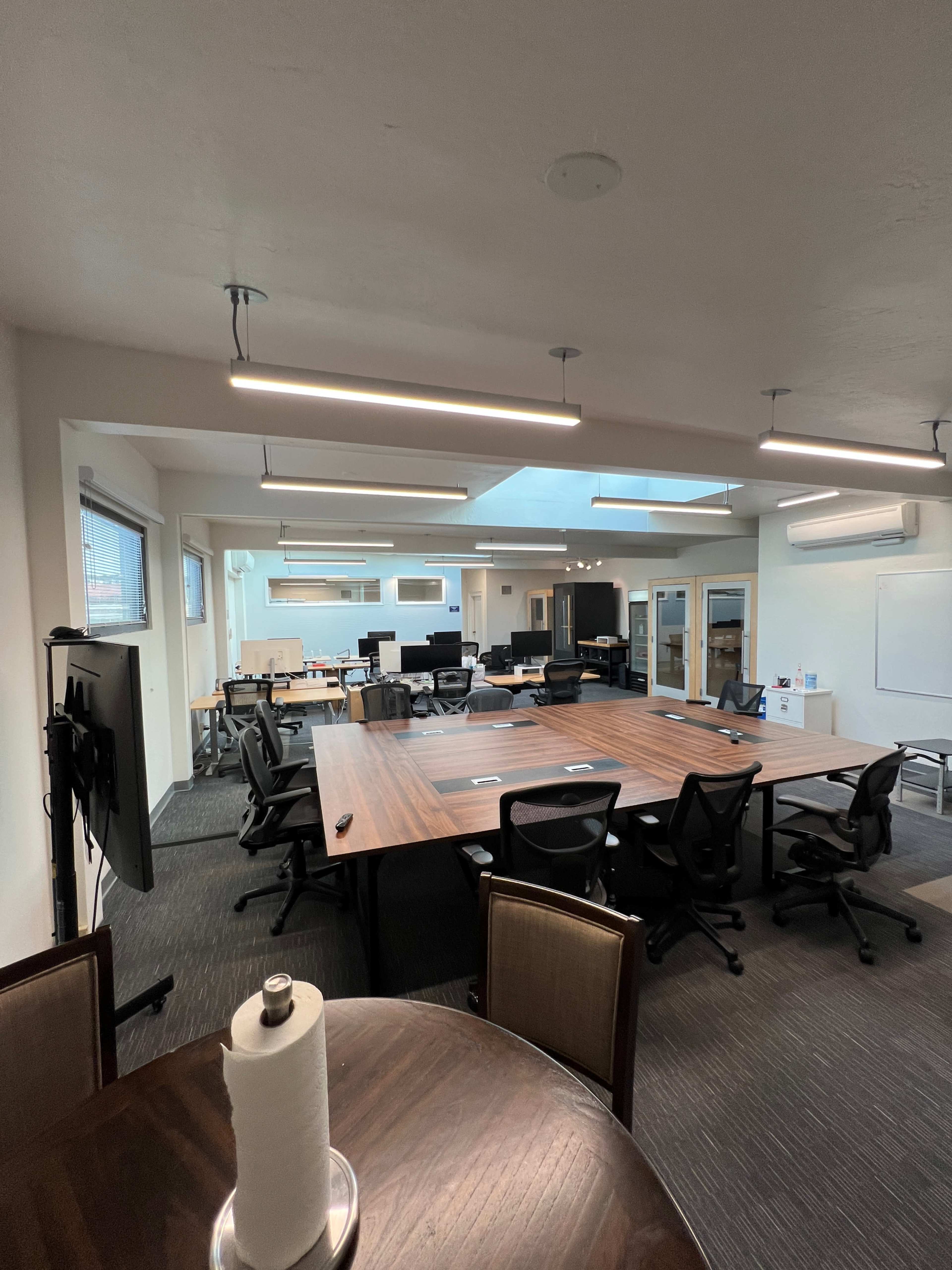 The image shows a modern office meeting room with a large wooden table surrounded by black chairs, multiple workstations in the background, and overhead lighting.