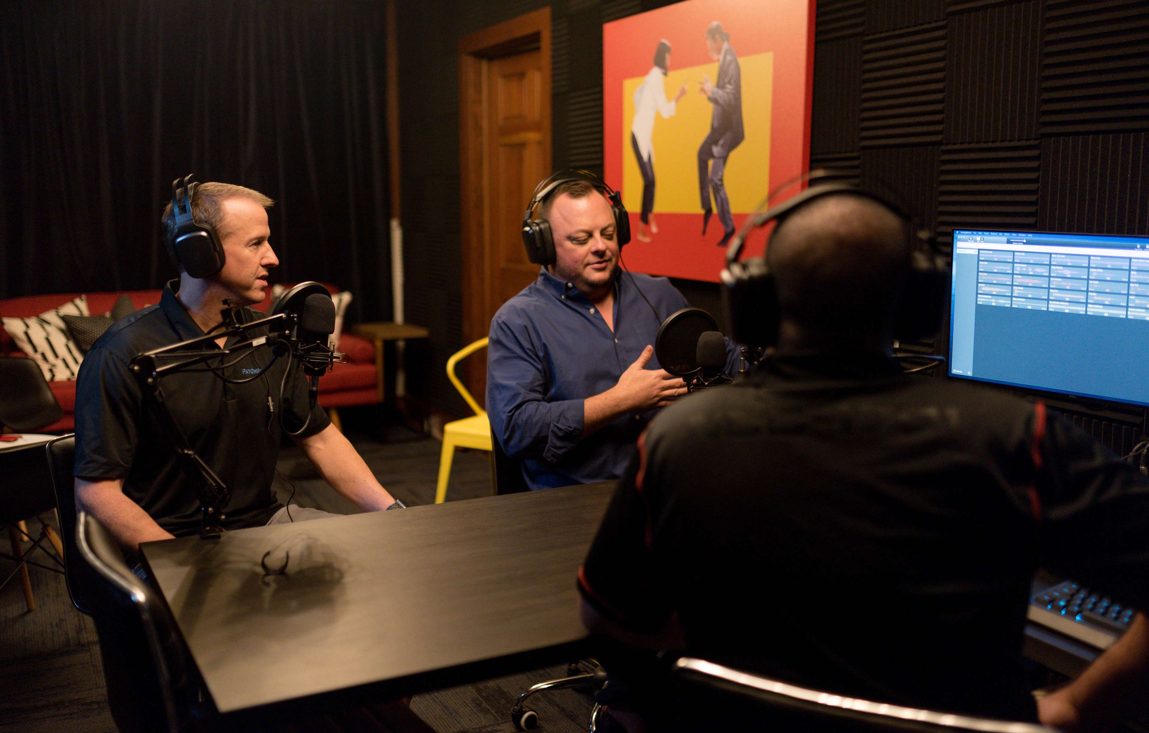 Three men sit at a table in a recording studio, each with a microphone in front of them, while one man speaks and another listens attentively.