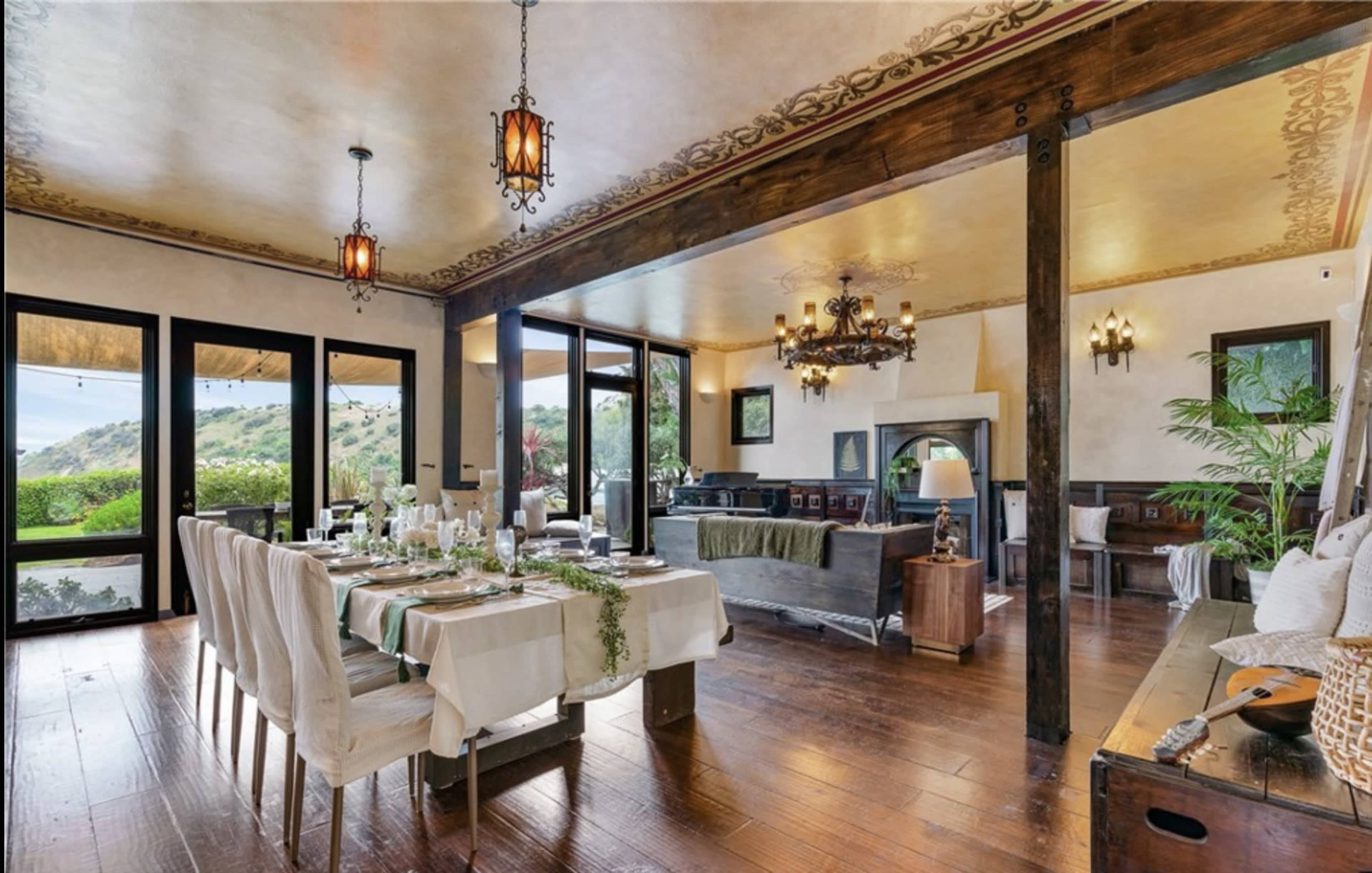 The image shows a spacious dining area with a long table set for a meal, surrounded by large windows that overlook a scenic view.