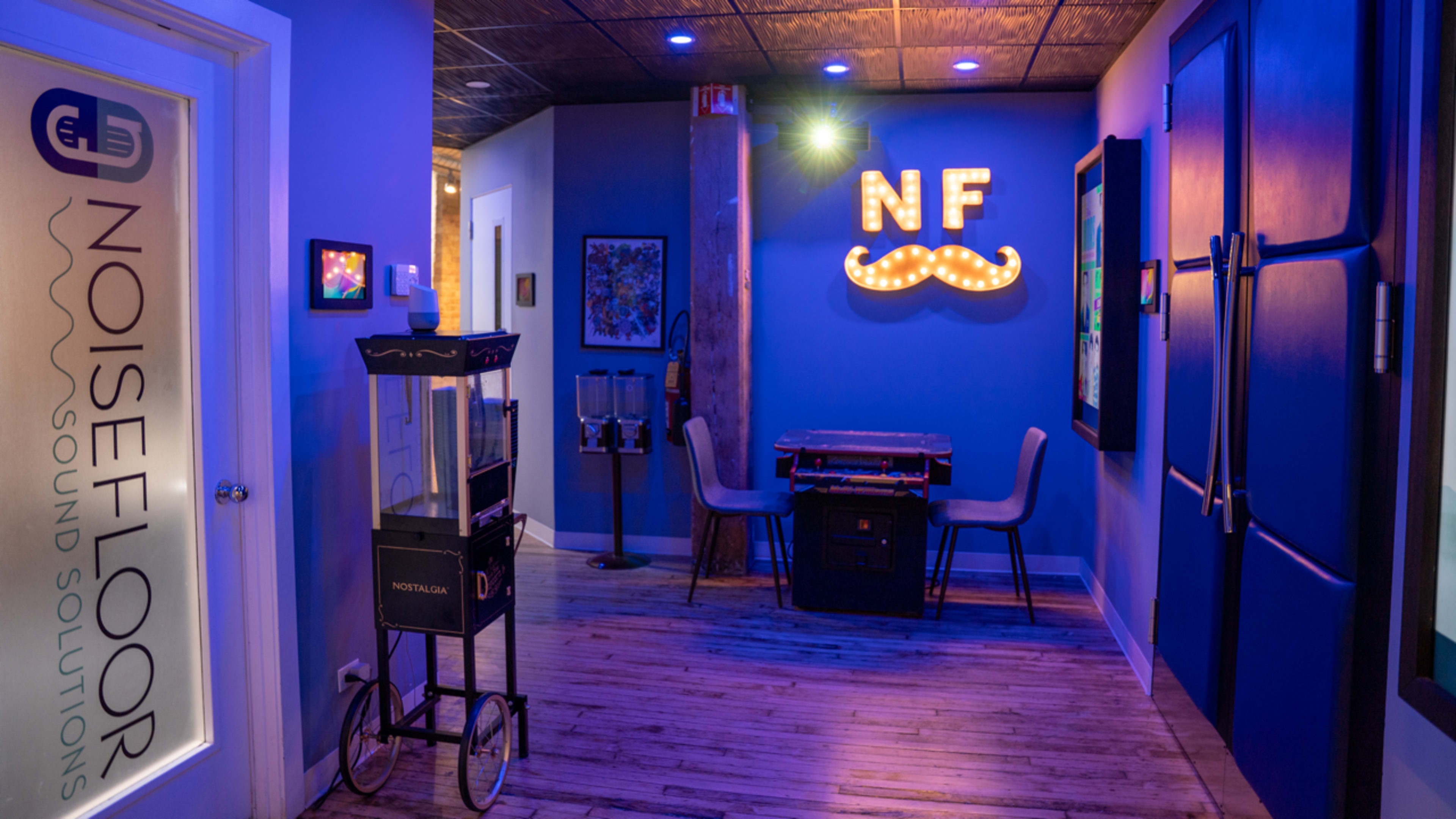 The image shows a hallway with a glass door marked "NOISEFLOOR SOUND SOLUTIONS," a decorated wall with a lit sign reading "NF," and a small cart beside a table and chairs.