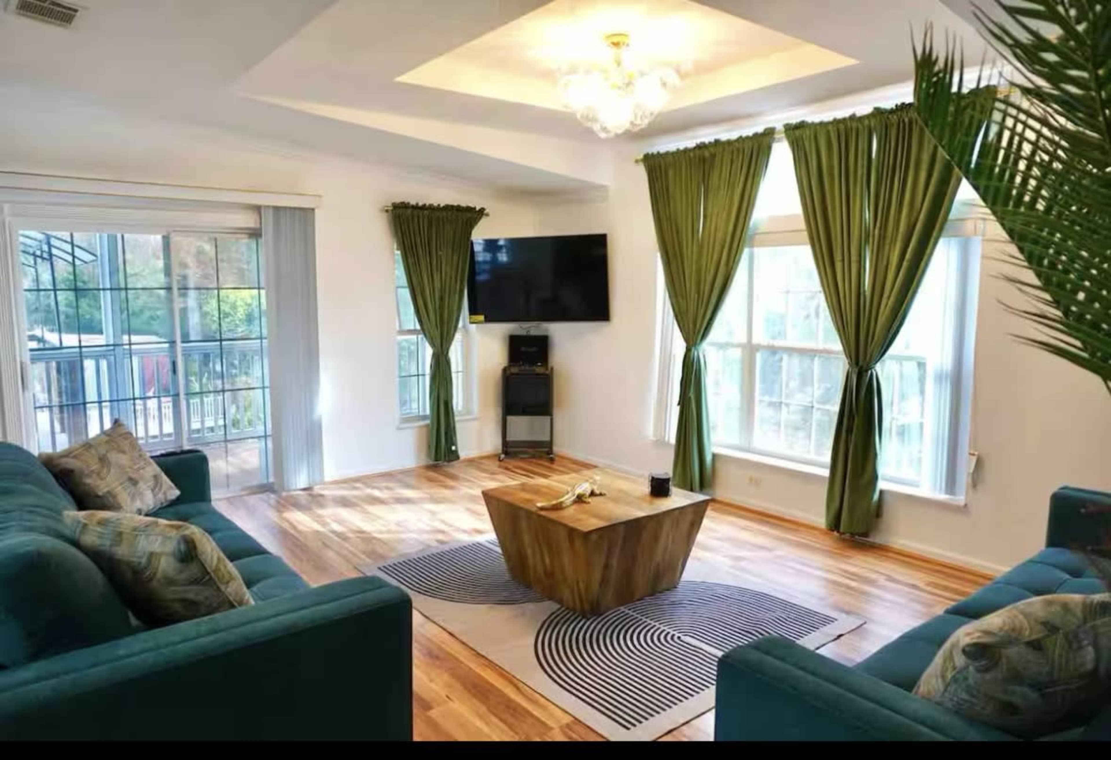 The living room features two green sofas facing a wooden coffee table, with large windows dressed in green curtains and a flat-screen TV mounted on the wall.
