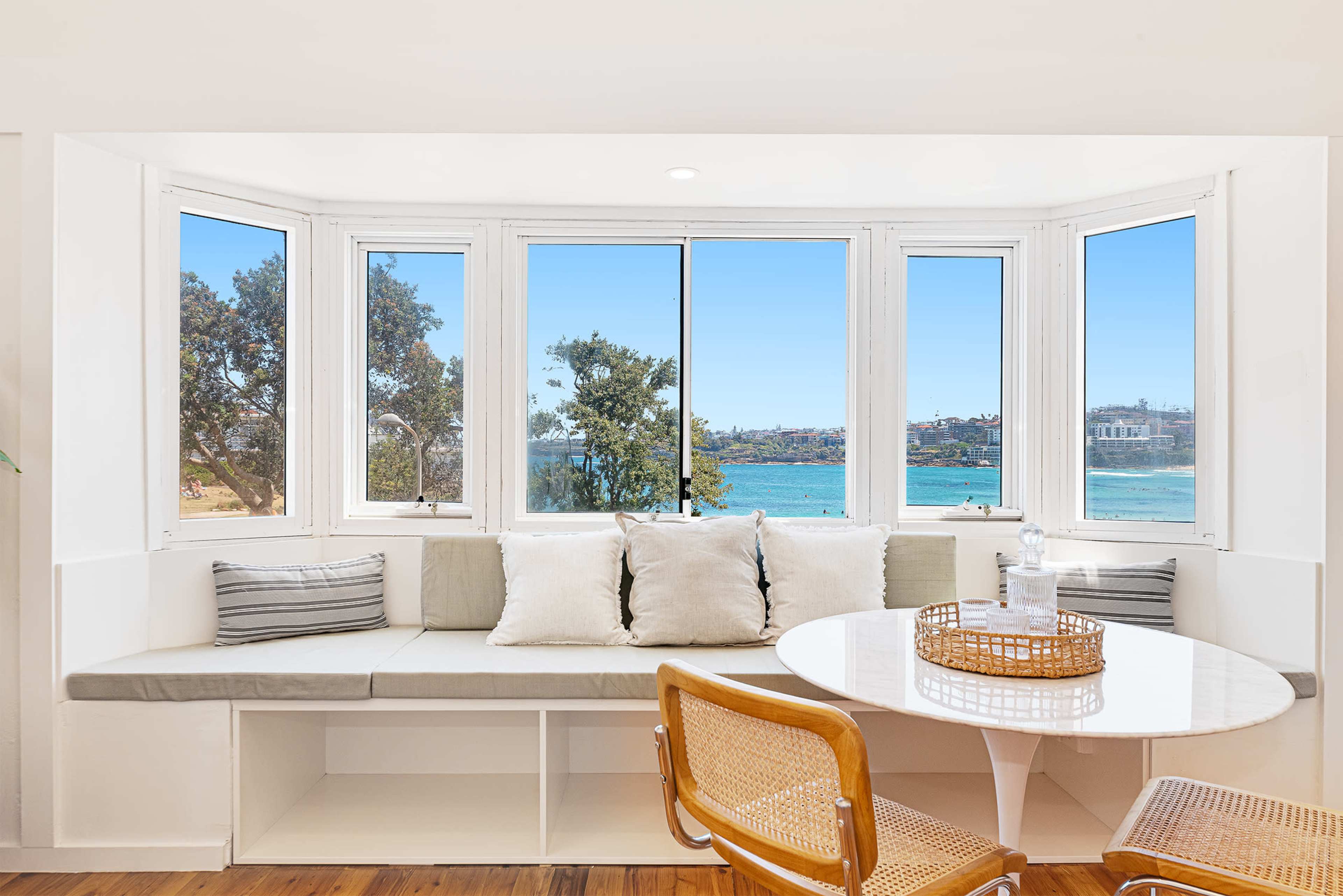A bright, modern seating area features a bay window with views of a beach and sea, complemented by a round dining table and stylish chairs.