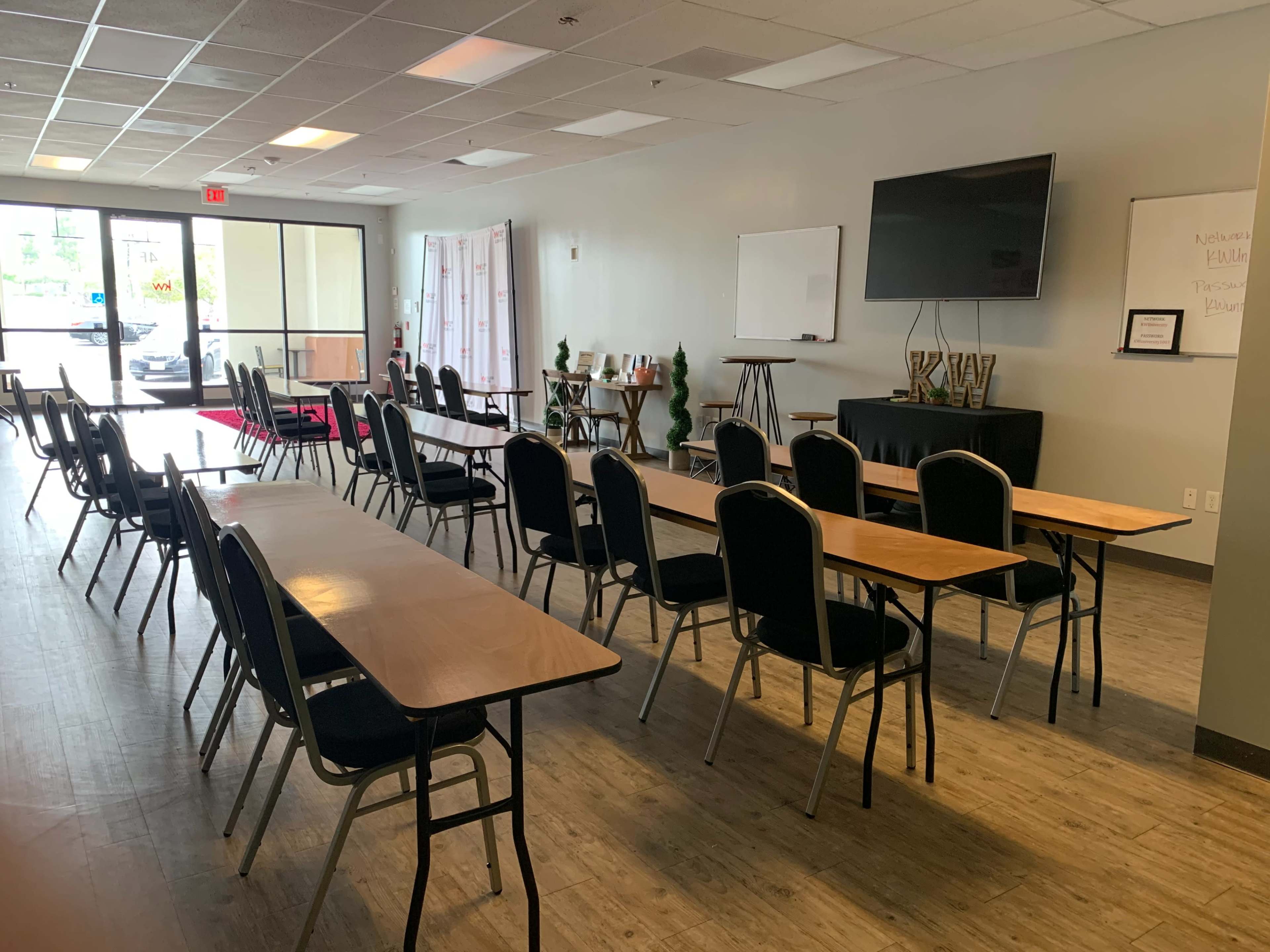 An interior room set up with multiple rows of tables and chairs, a large television, and a whiteboard at the front.