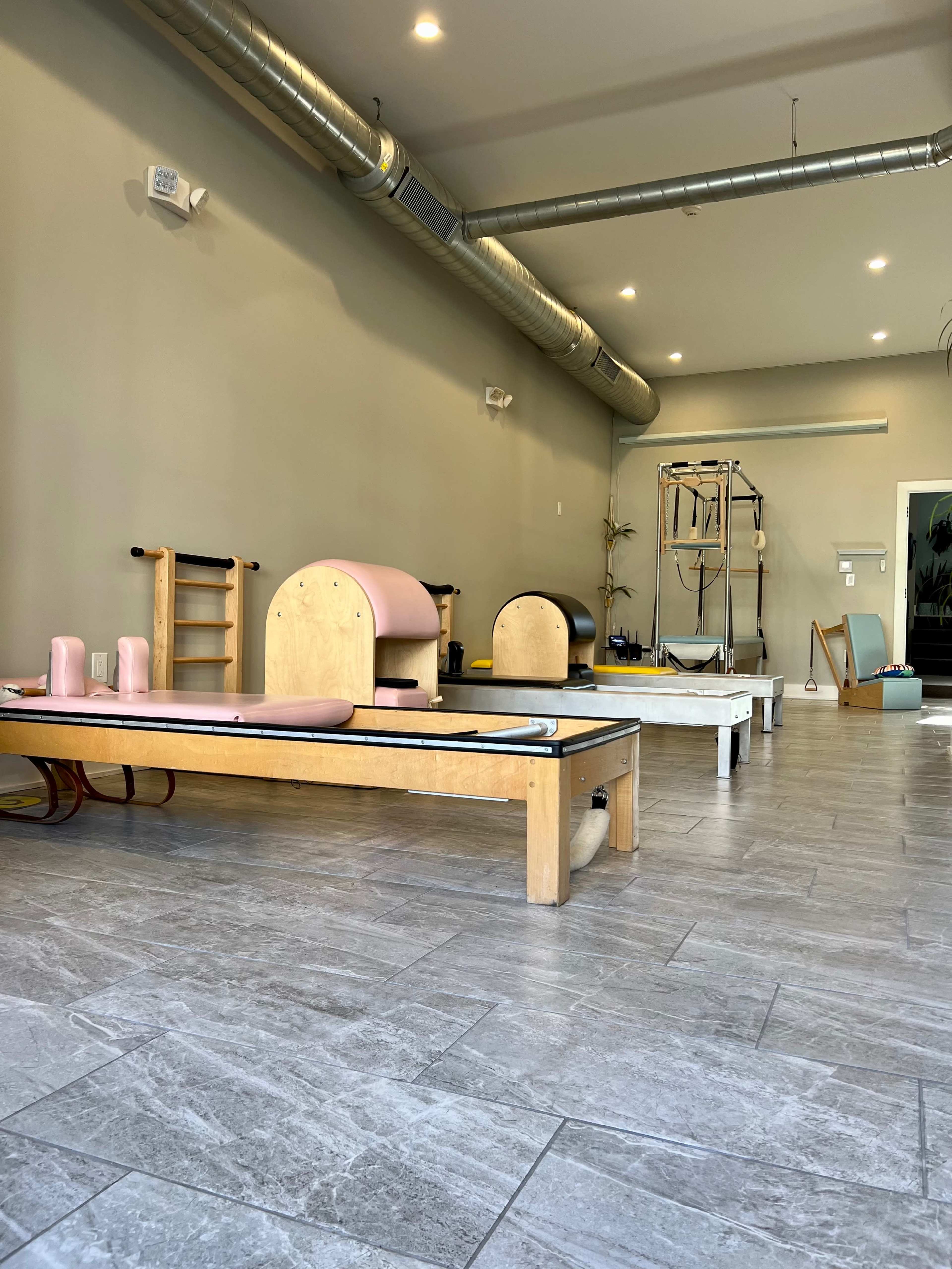 A spacious Pilates studio with wooden reformers and equipment arranged neatly on gray tiled flooring.
