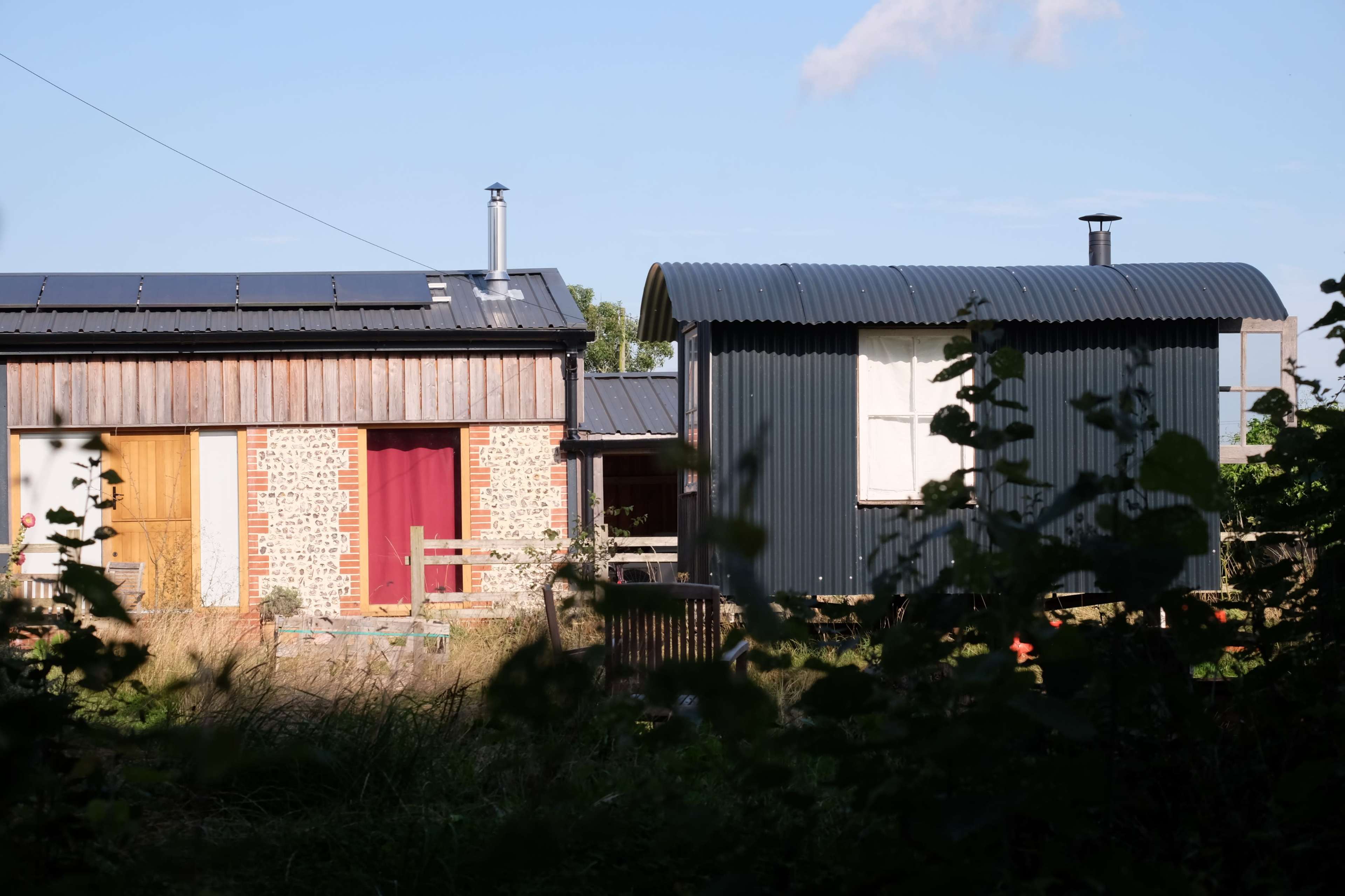 The image shows a modern wooden house with a corrugated metal extension, partially obscured by tall grass and foliage.
