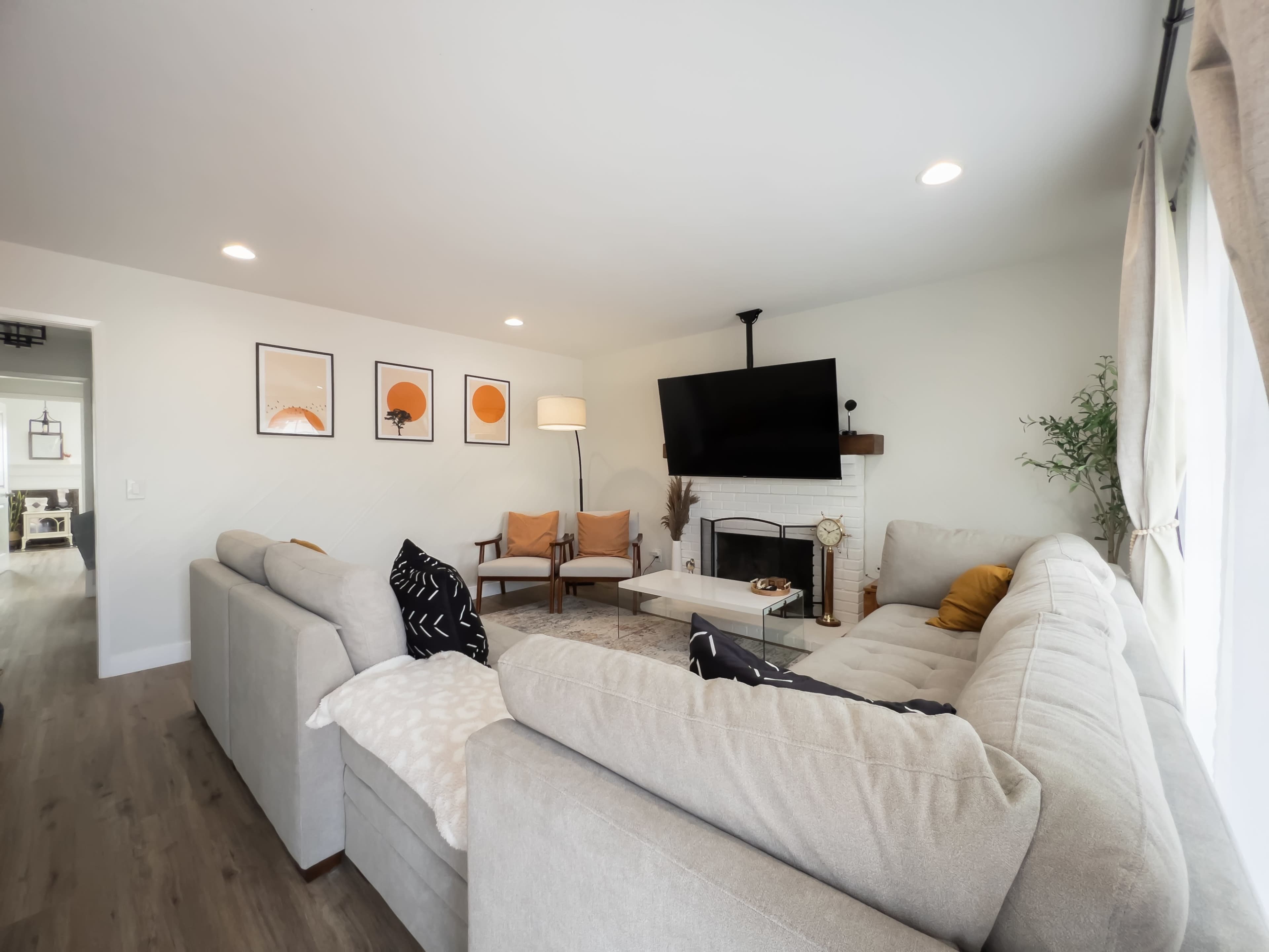 A modern living room features a large sectional sofa, a coffee table, and artwork on the walls, with a TV mounted above a fireplace.