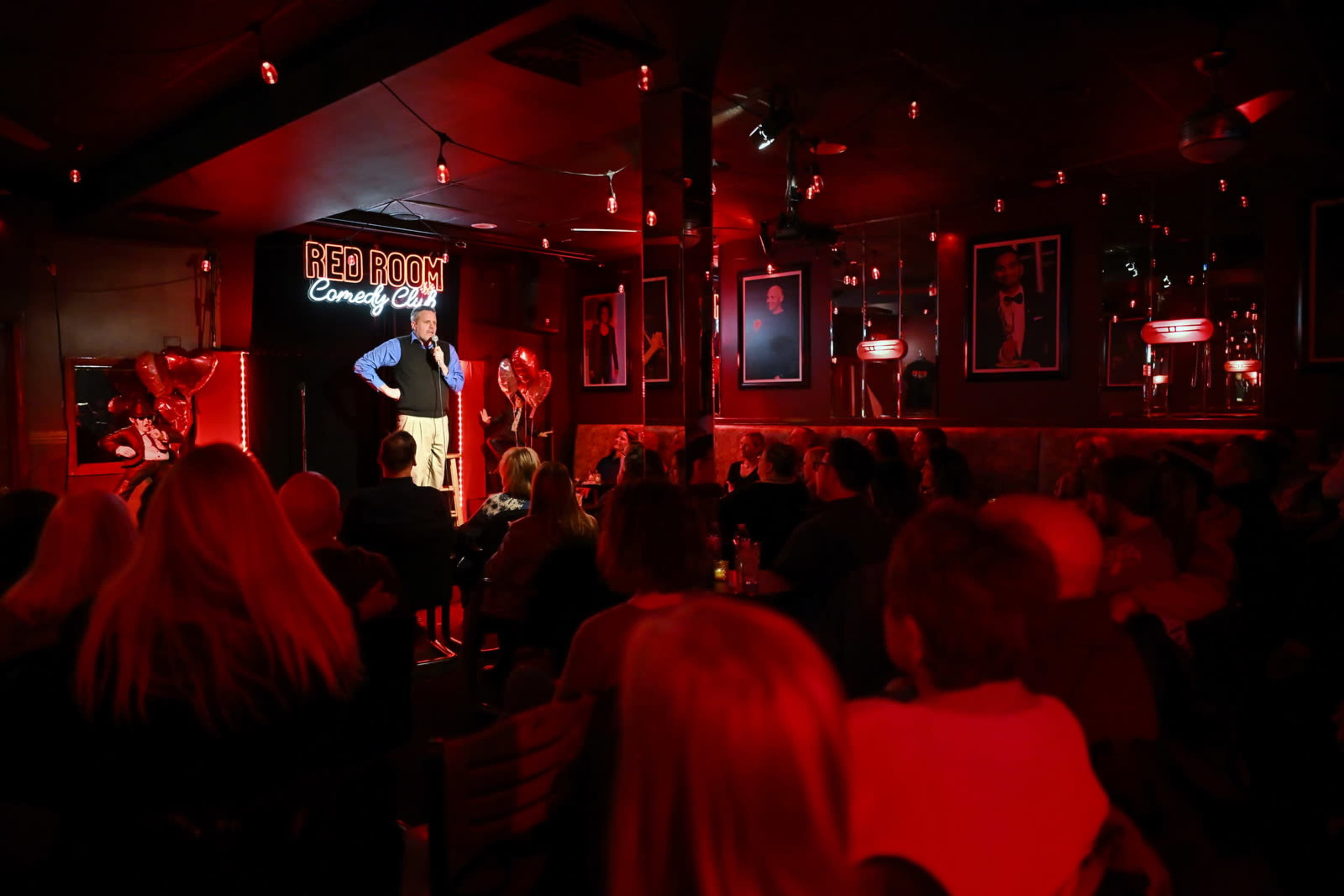 A comedian performs on stage at the Red Room Comedy Club while an audience watches, illuminated by red lighting.