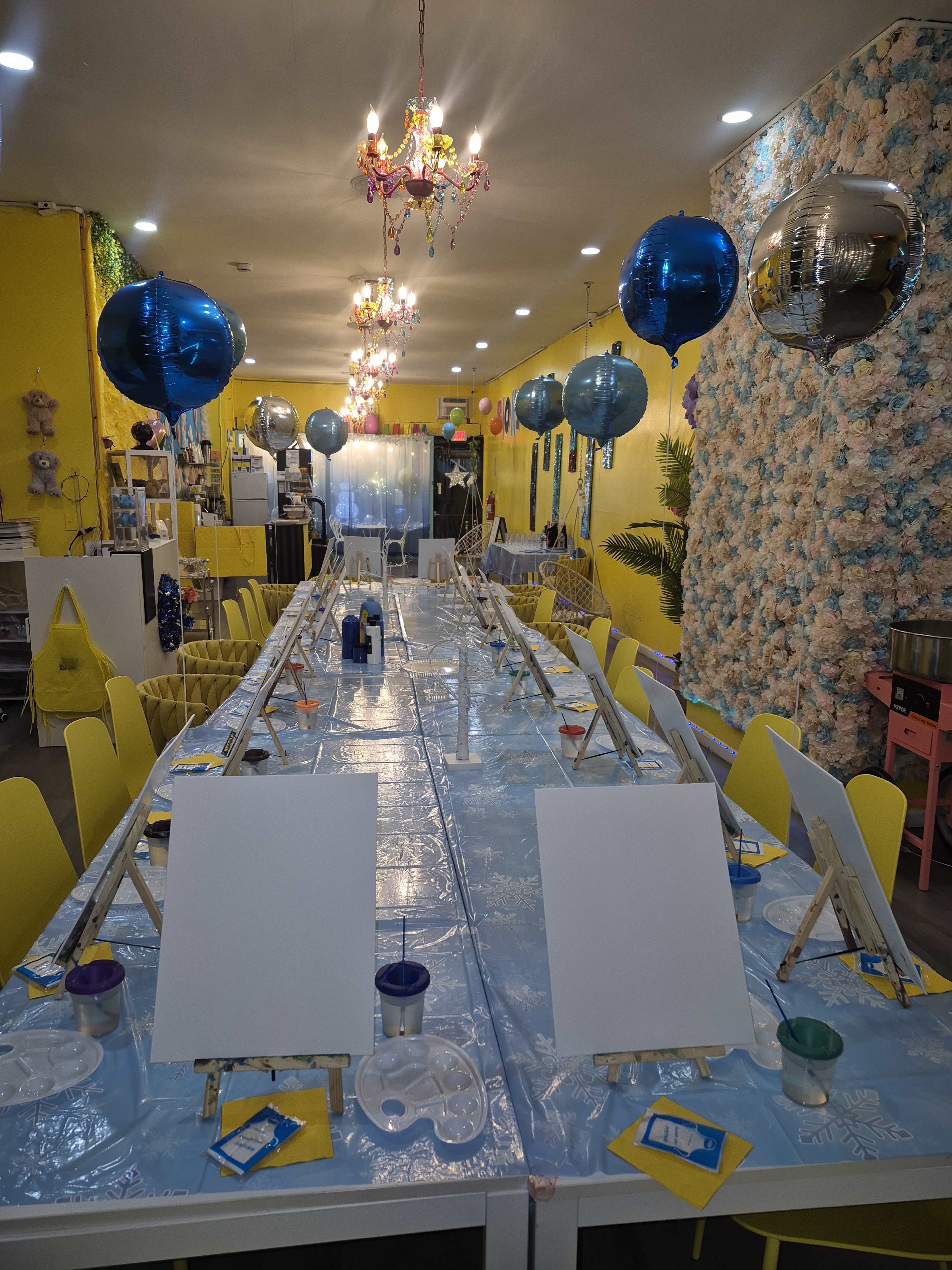 The image shows a brightly decorated art studio with a long table set for a painting activity, featuring blank canvases and pastel-colored supplies arranged on a blue tablecloth.