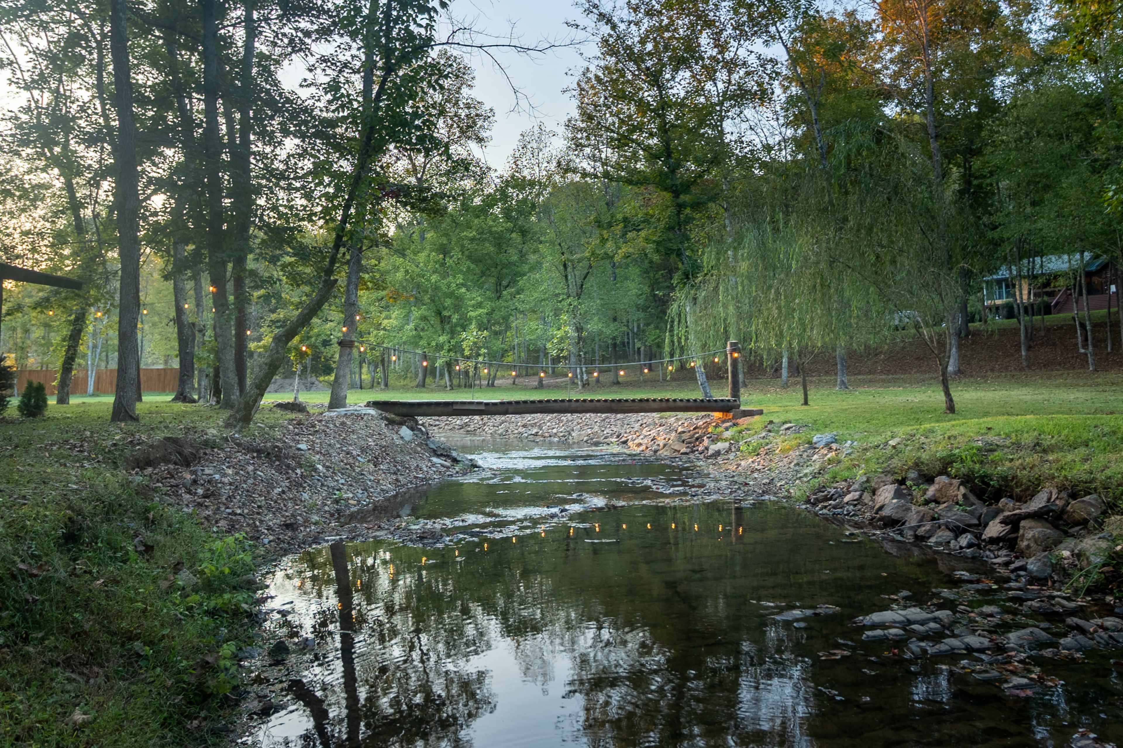 A wooden bridge crosses a shallow creek surrounded by trees and soft grass, with string lights illuminating the area.