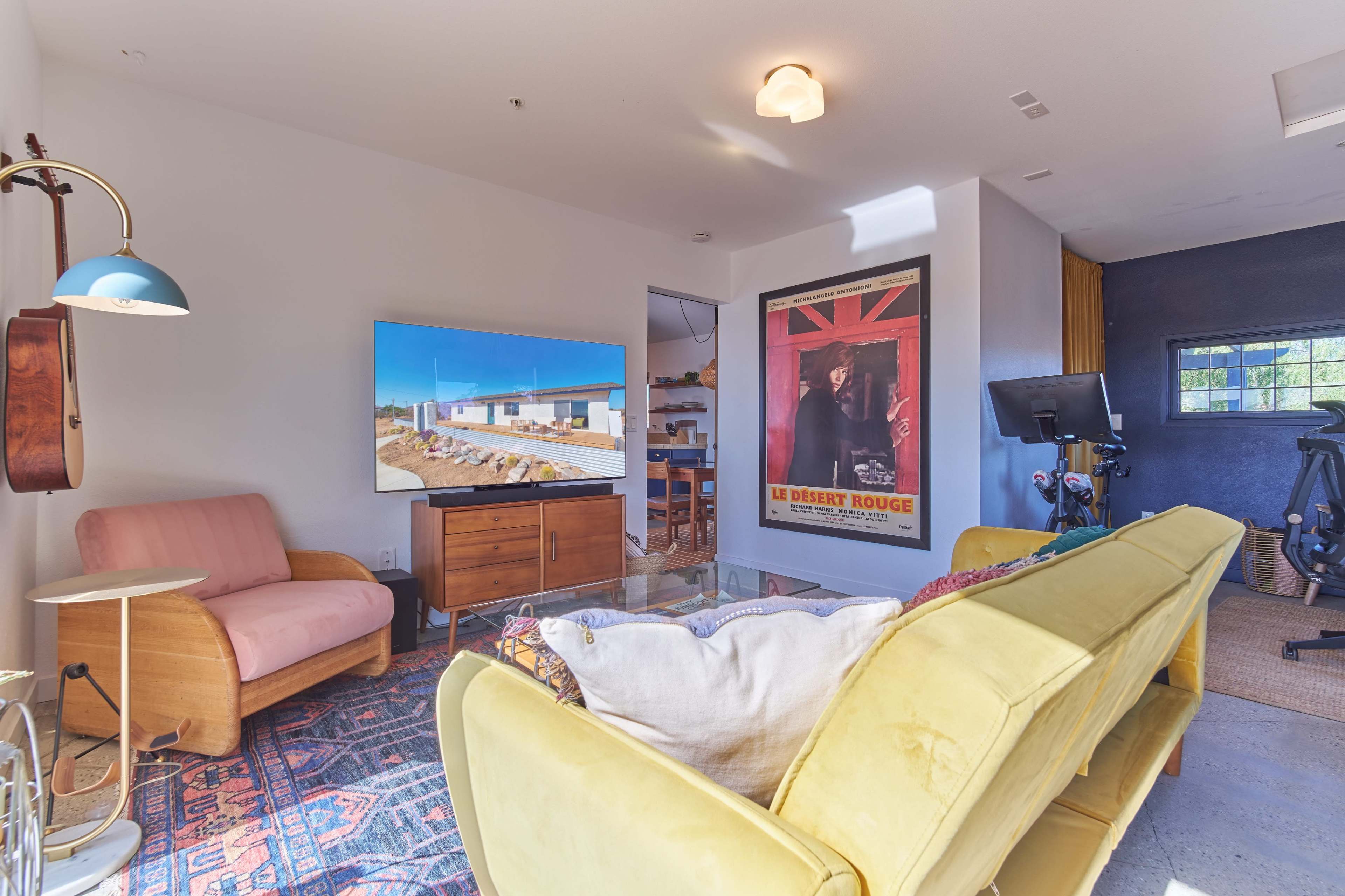 The living room features a yellow couch, a pink armchair, a large television displaying an exterior view of a modern house, and a wall decorated with a framed poster.