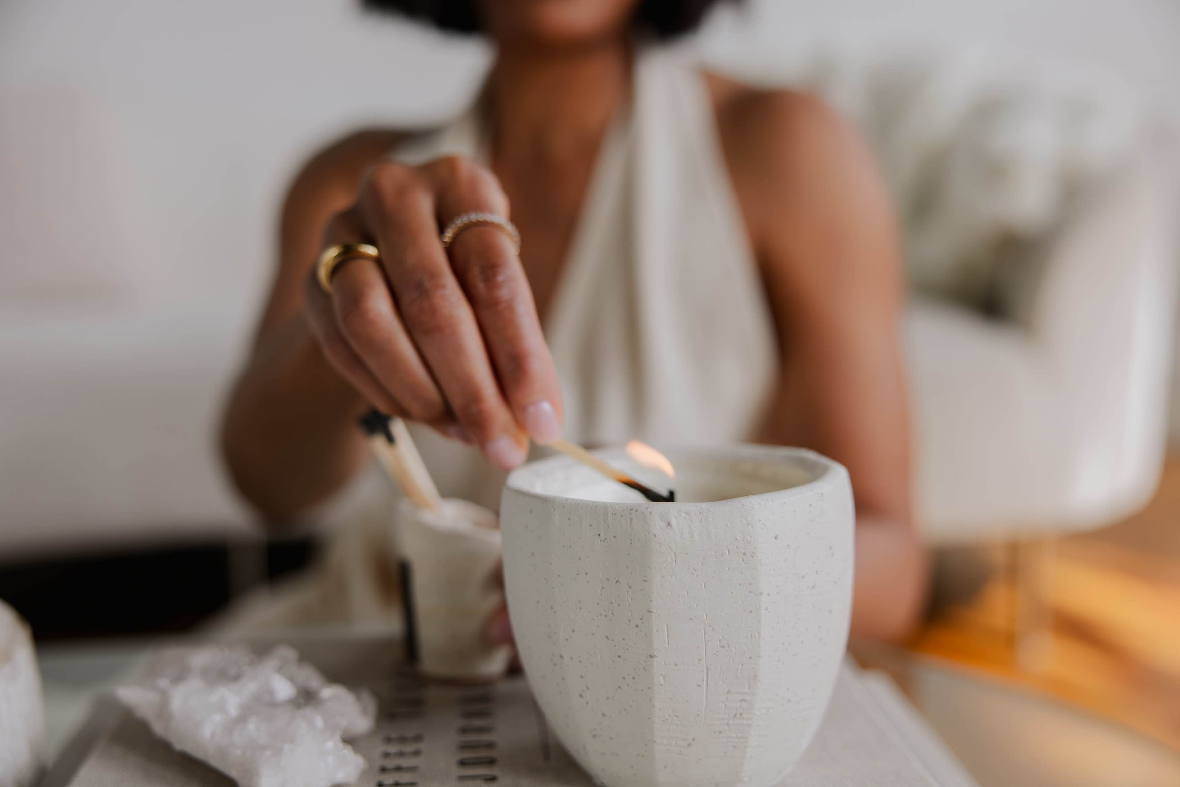 A person lights a candle in a textured white pot using a match, with decorative crystals nearby.