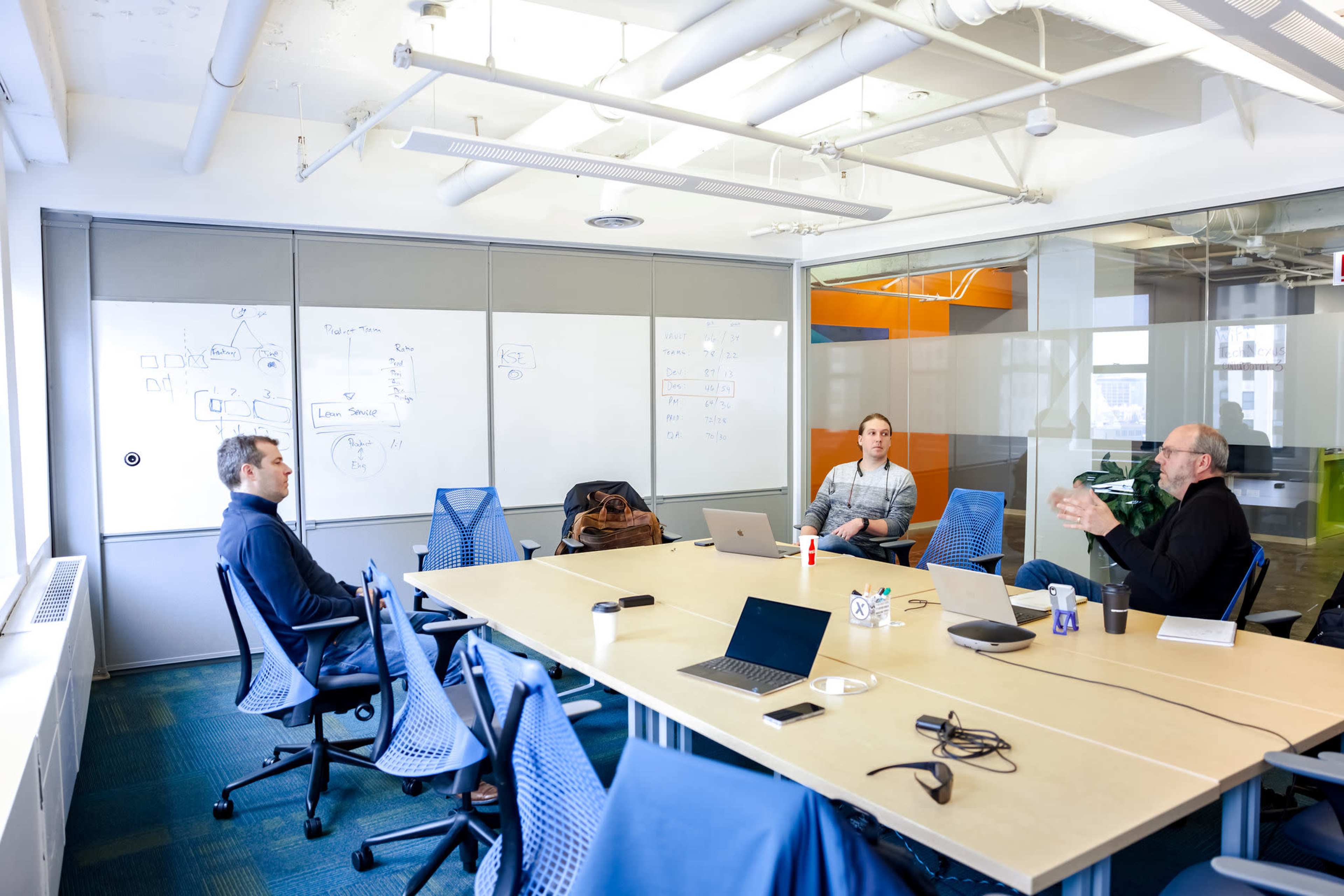 Three people are seated around a conference table in a bright meeting room with a whiteboard and laptops.