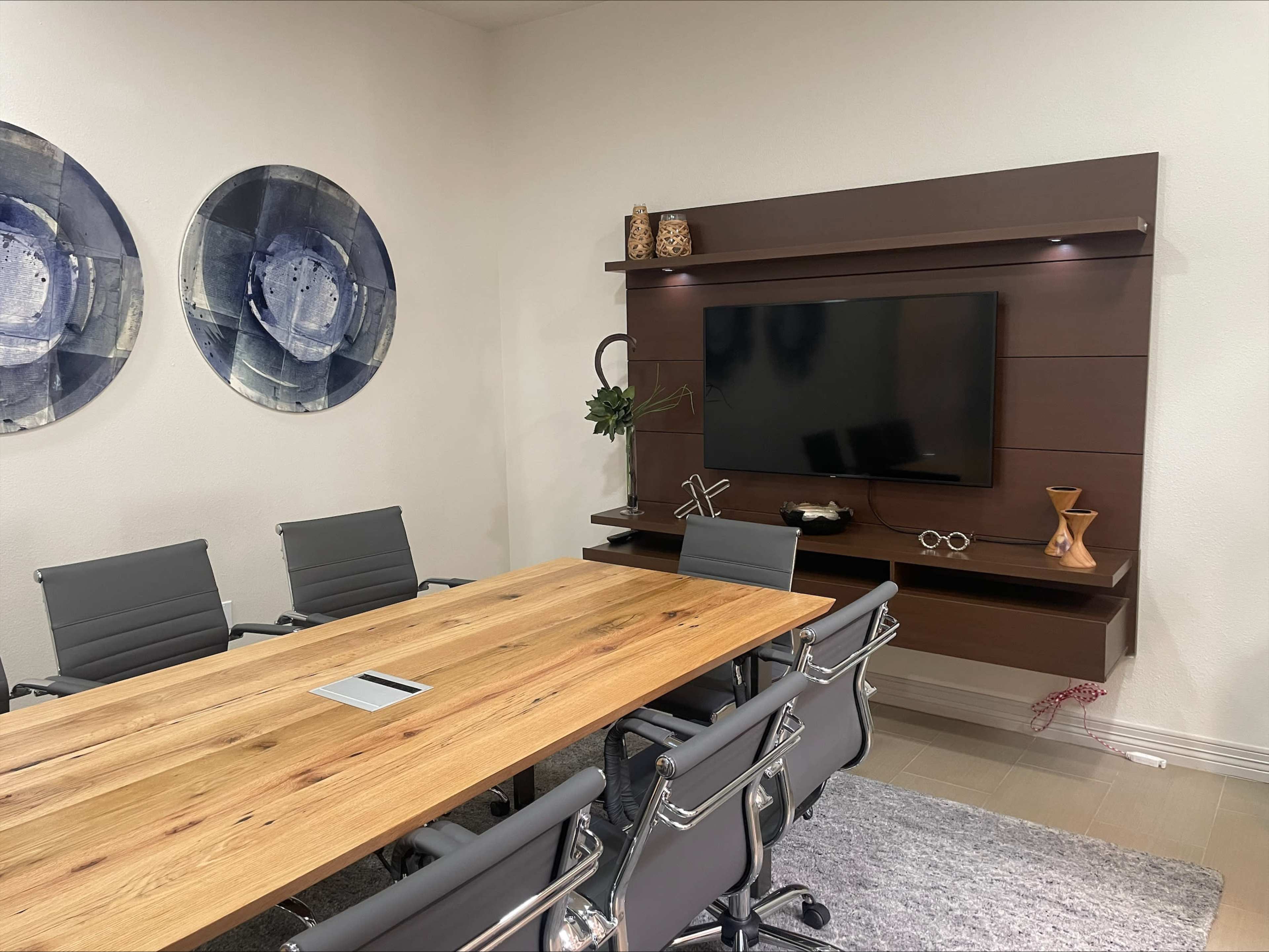 A modern conference room features a large wooden table, gray ergonomic chairs, and a wall-mounted television surrounded by decorative items.