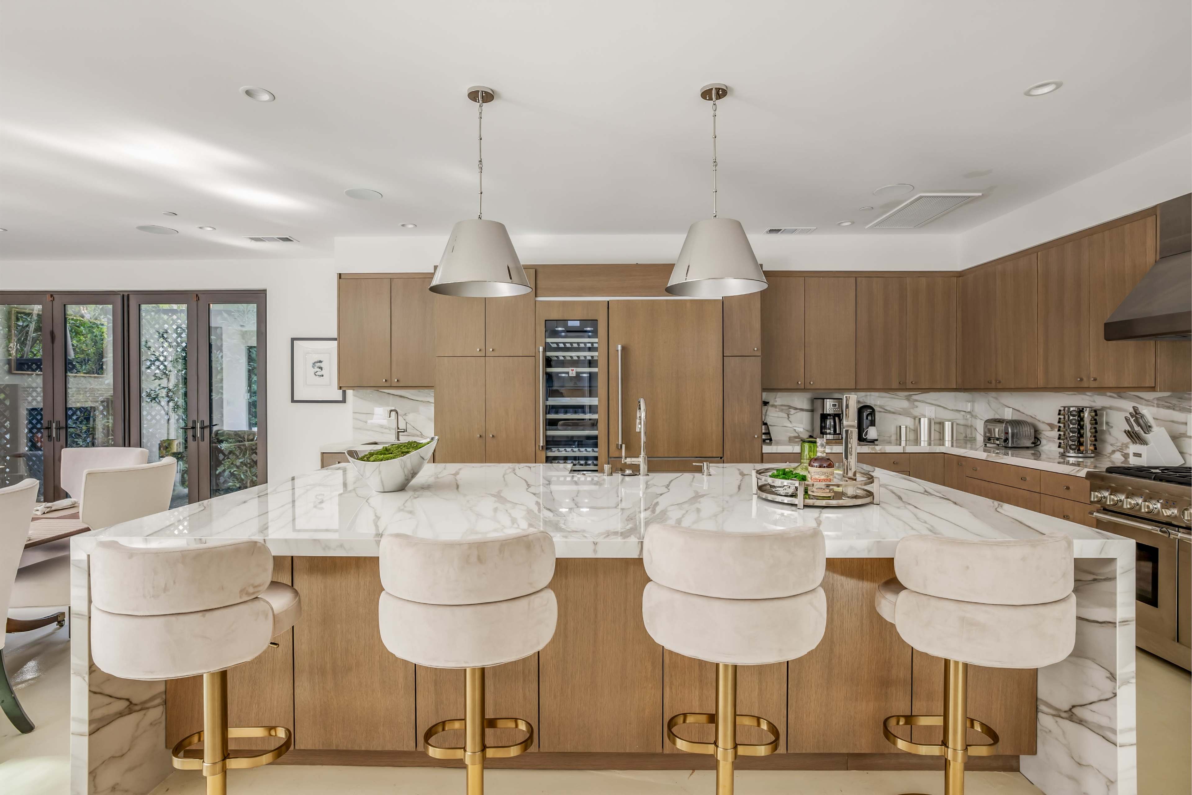The image shows a modern kitchen with a marble countertop island, four bar stools, wooden cabinetry, and stainless steel appliances.