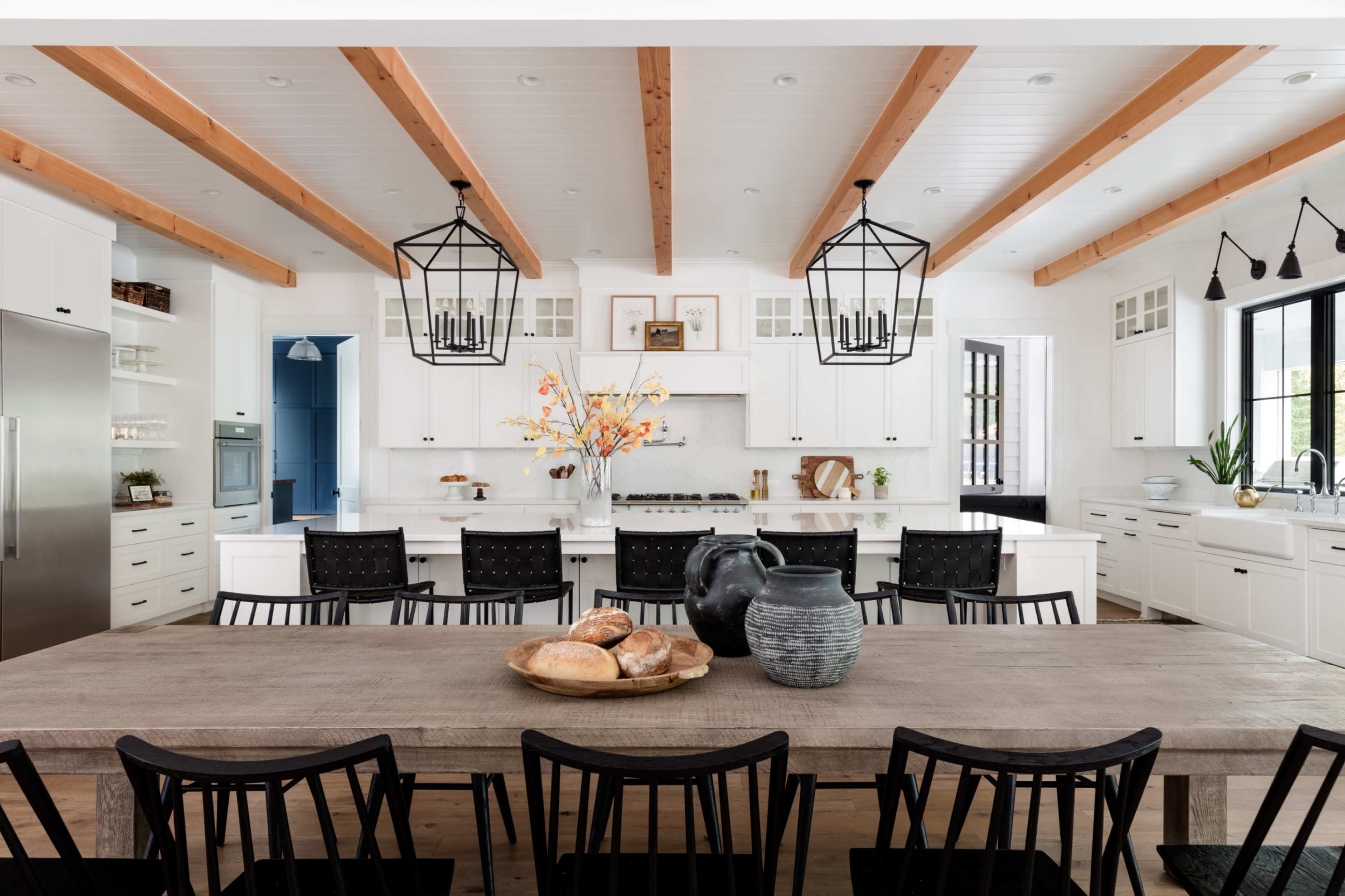 A spacious kitchen features a long wooden dining table surrounded by black chairs, with white cabinetry and exposed wooden beams overhead.