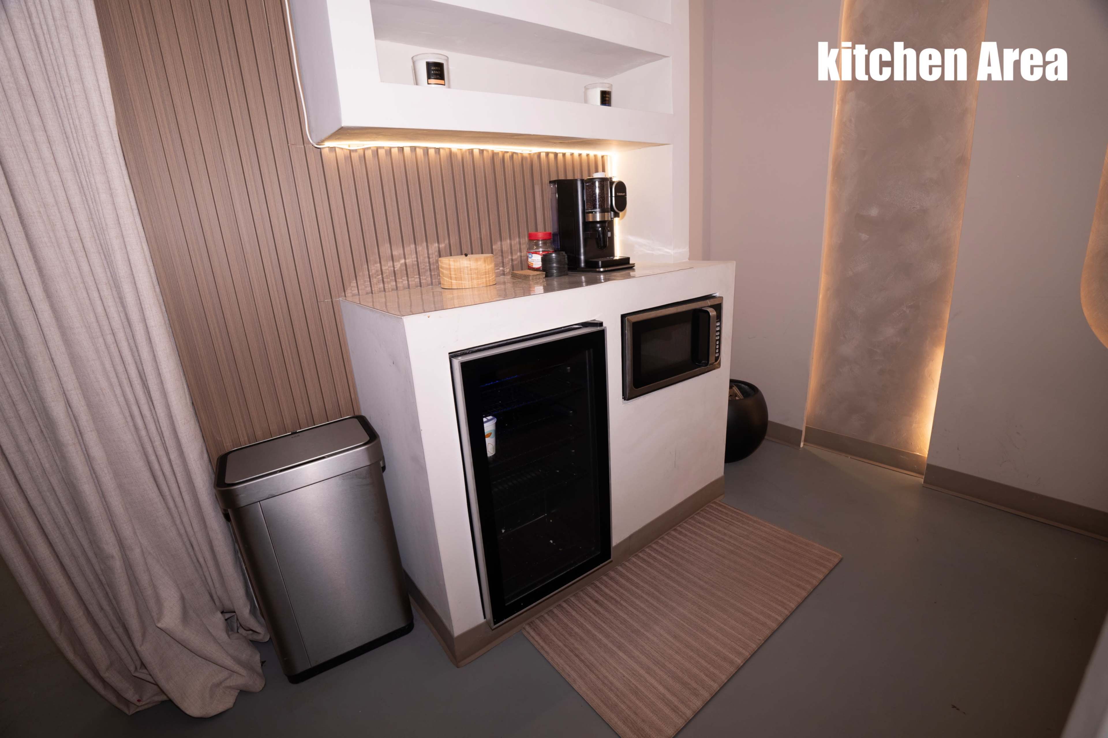 The image depicts a compact kitchen area featuring a coffee maker, a microwave, a mini fridge, a trash can, and a countertop with ambient lighting.