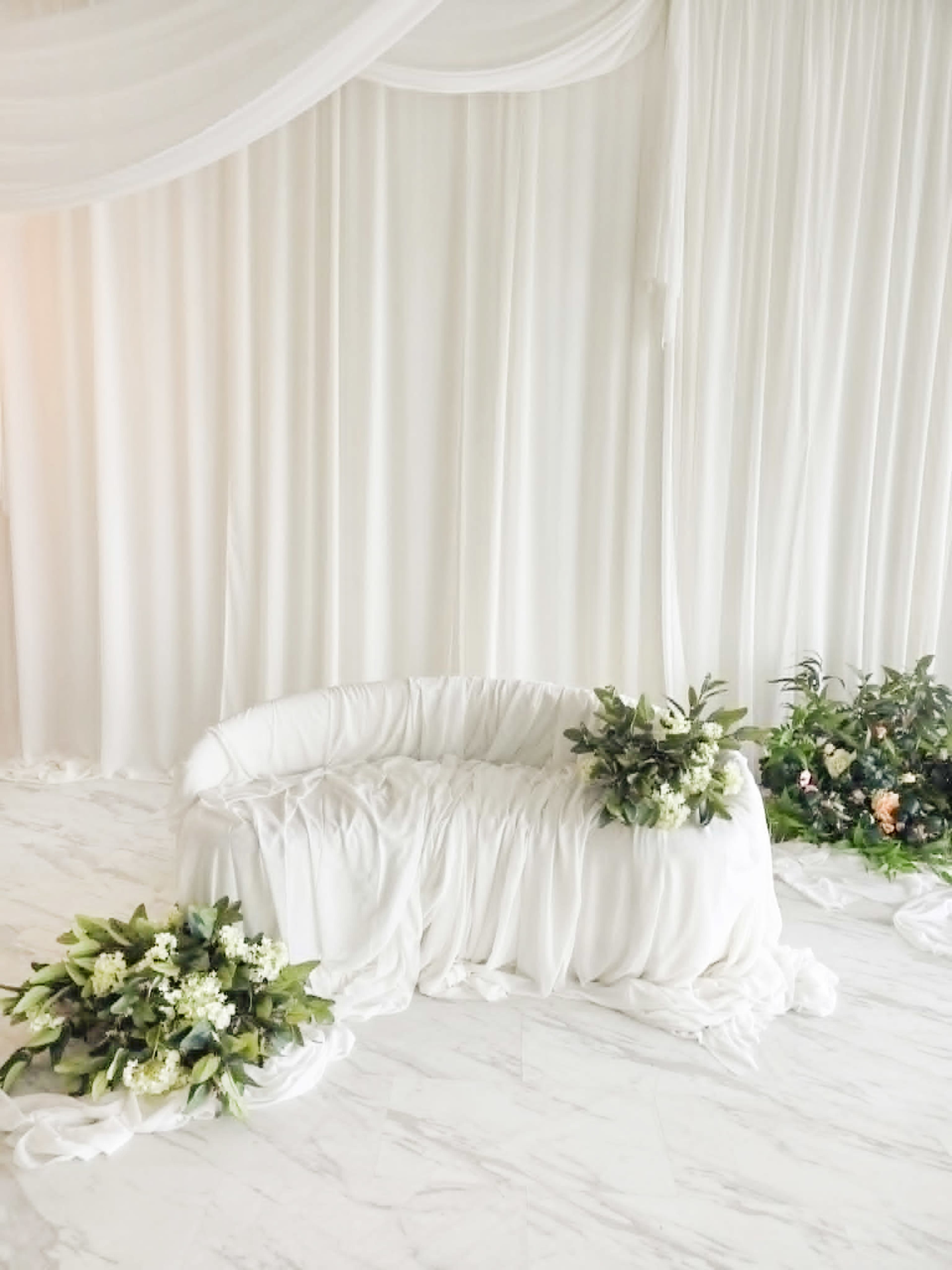 The scene features a white draped backdrop with a curved white sofa and floral arrangements on either side.