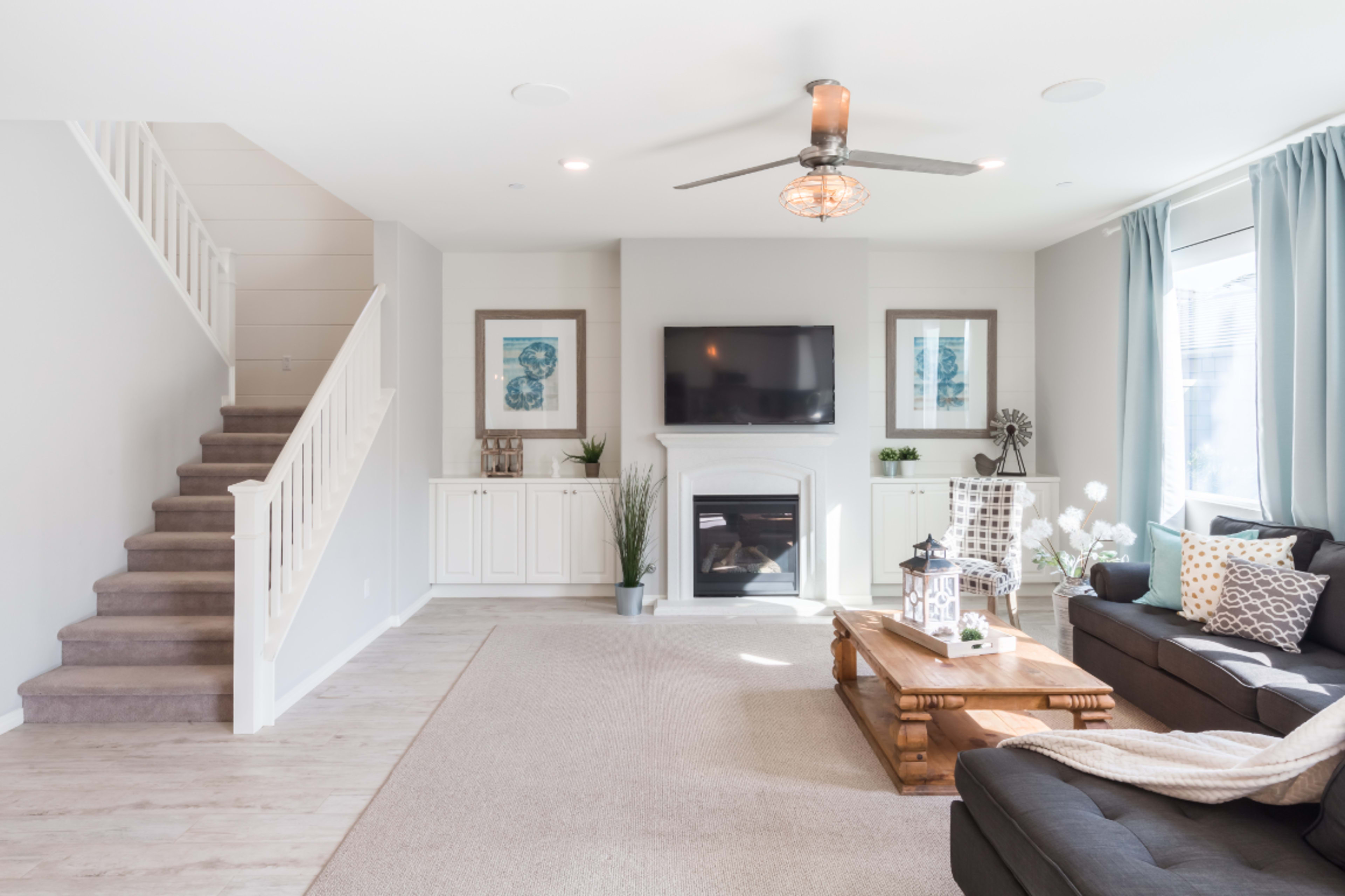 A modern living room features a gray couch, a wooden coffee table, and a staircase, with a television mounted above a white fireplace and light blue curtains framing a window.